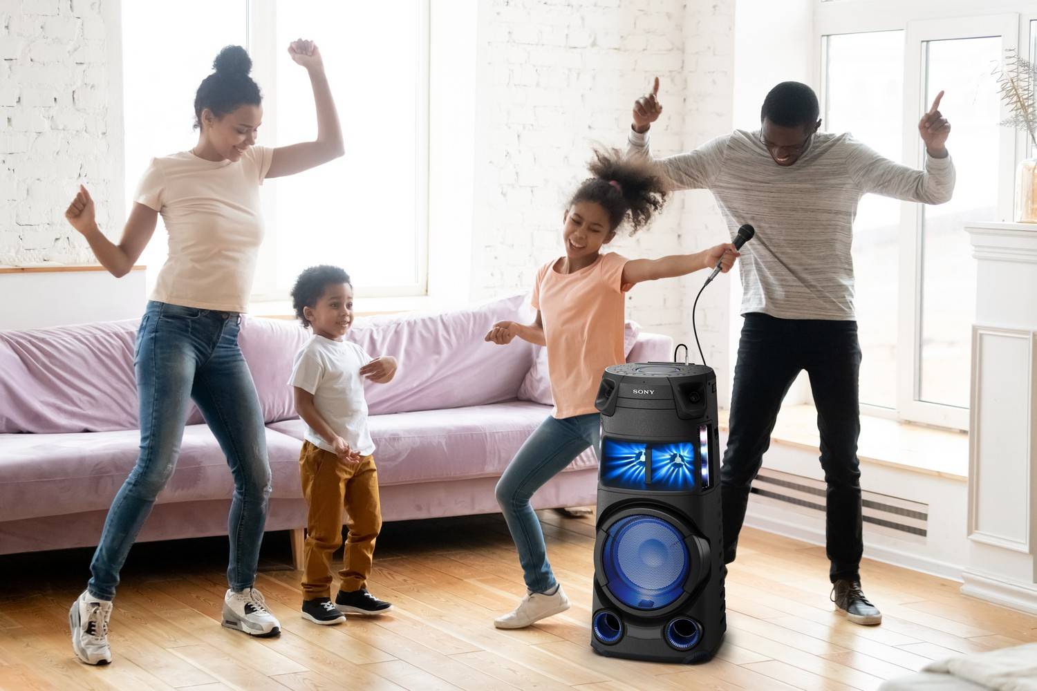 La familia baila alrededor de un altavoz. Una niña sostiene un micrófono. Un niño está de pie. Un sofá en el fondo.