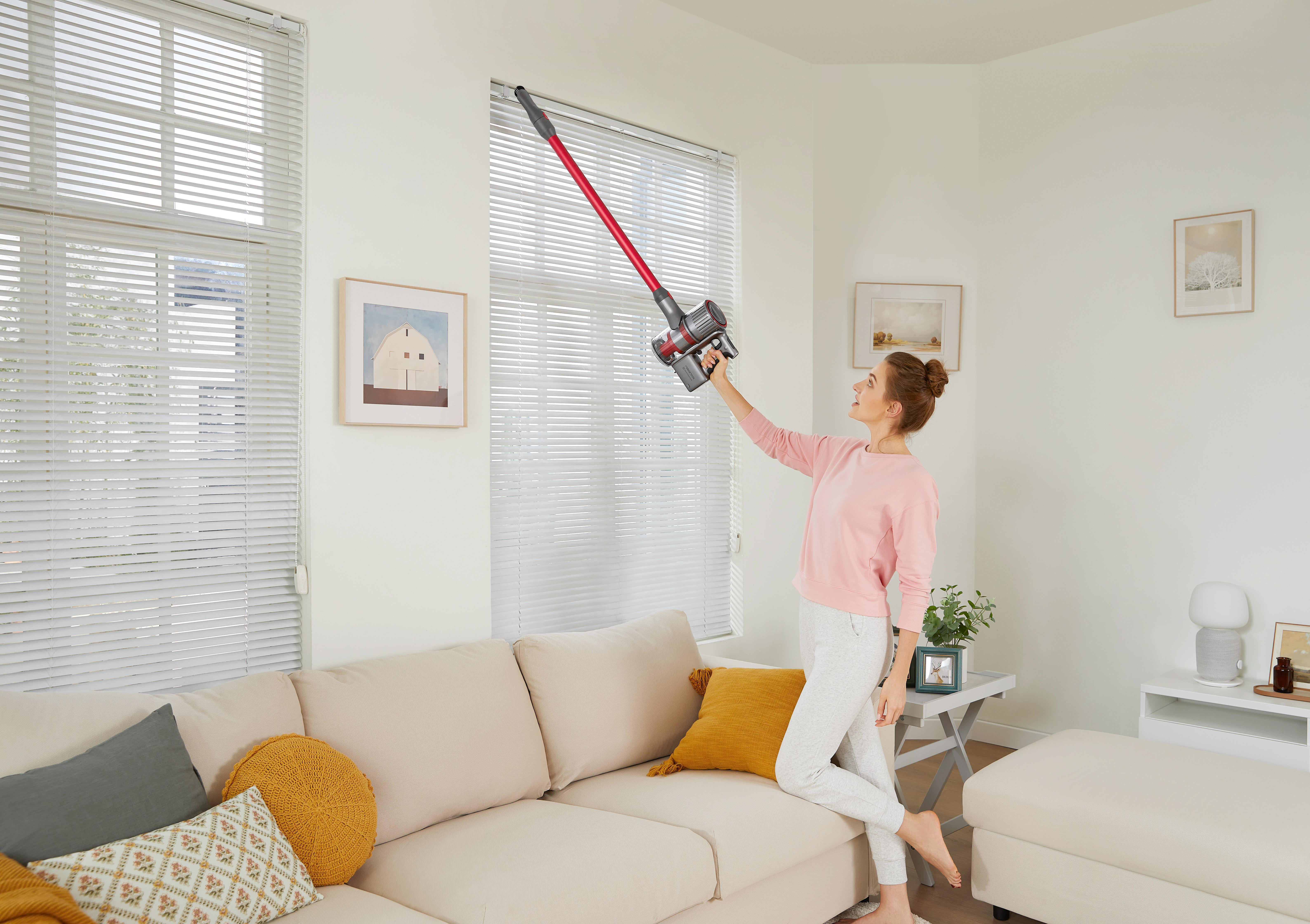 Frau, die ein Fenster saugt. Sie steht in der Nähe eines Sofas, die Jalousien sind sichtbar.