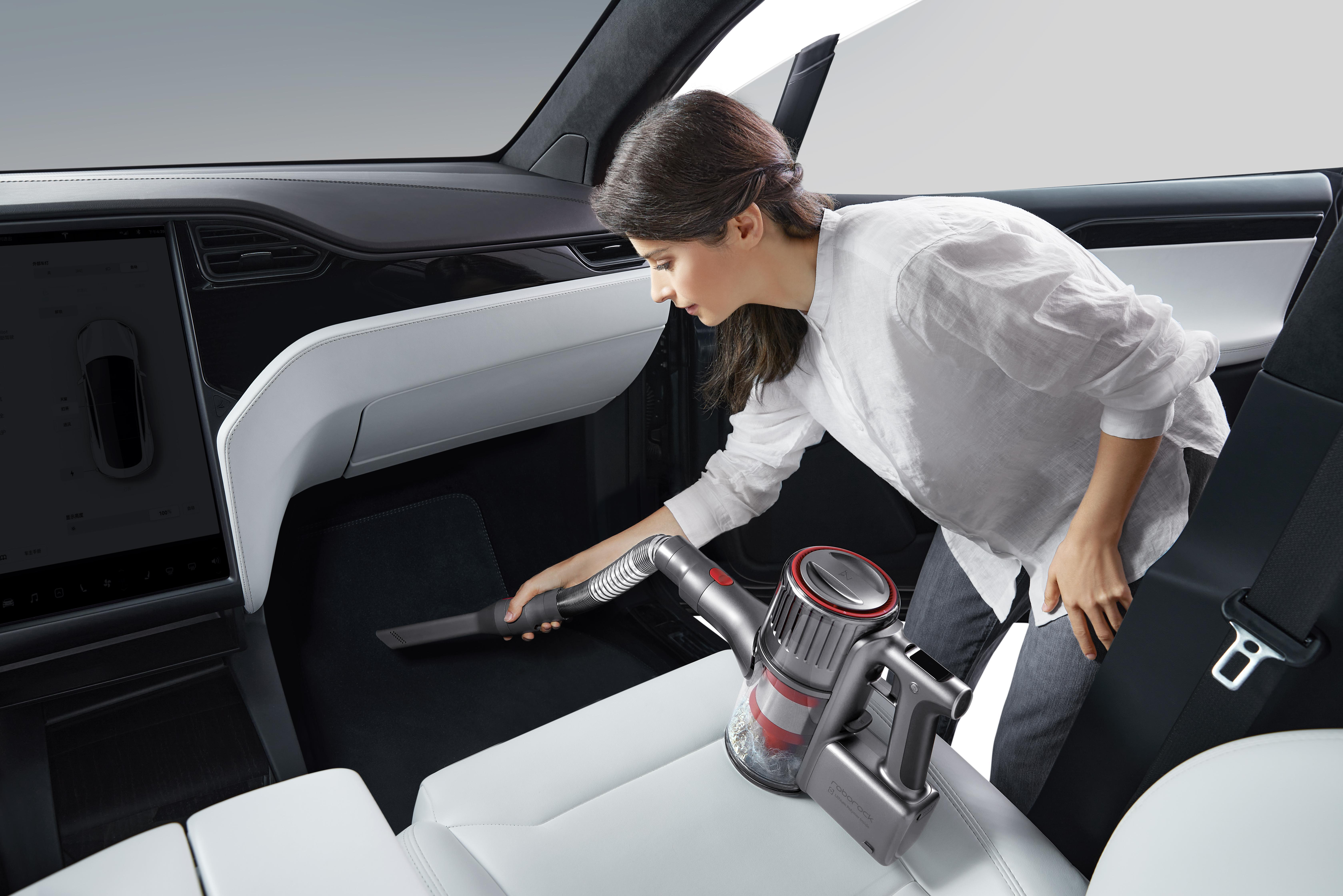 Frau saugt Innenraum eines Autos. Grauer Staubsauger, weiße Sitze, schwarzes Armaturenbrett. Das Auto steht in einer Garage.