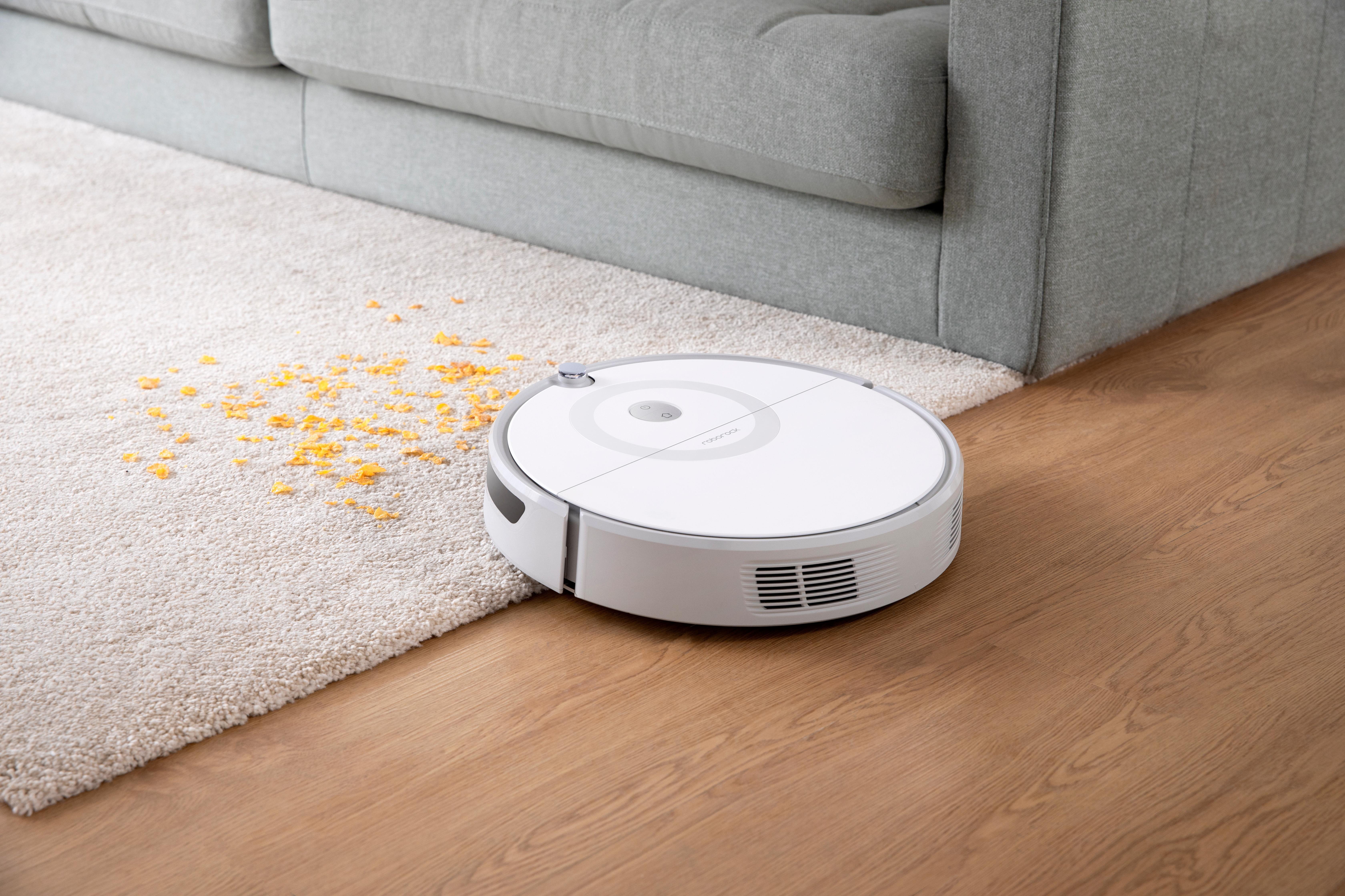 Ein weißer Roboter-Staubsauger bewegt sich auf einem Holzboden, in der Nähe eines Teppichs und eines grauen Sofas, mit verstreuten Trümmern.