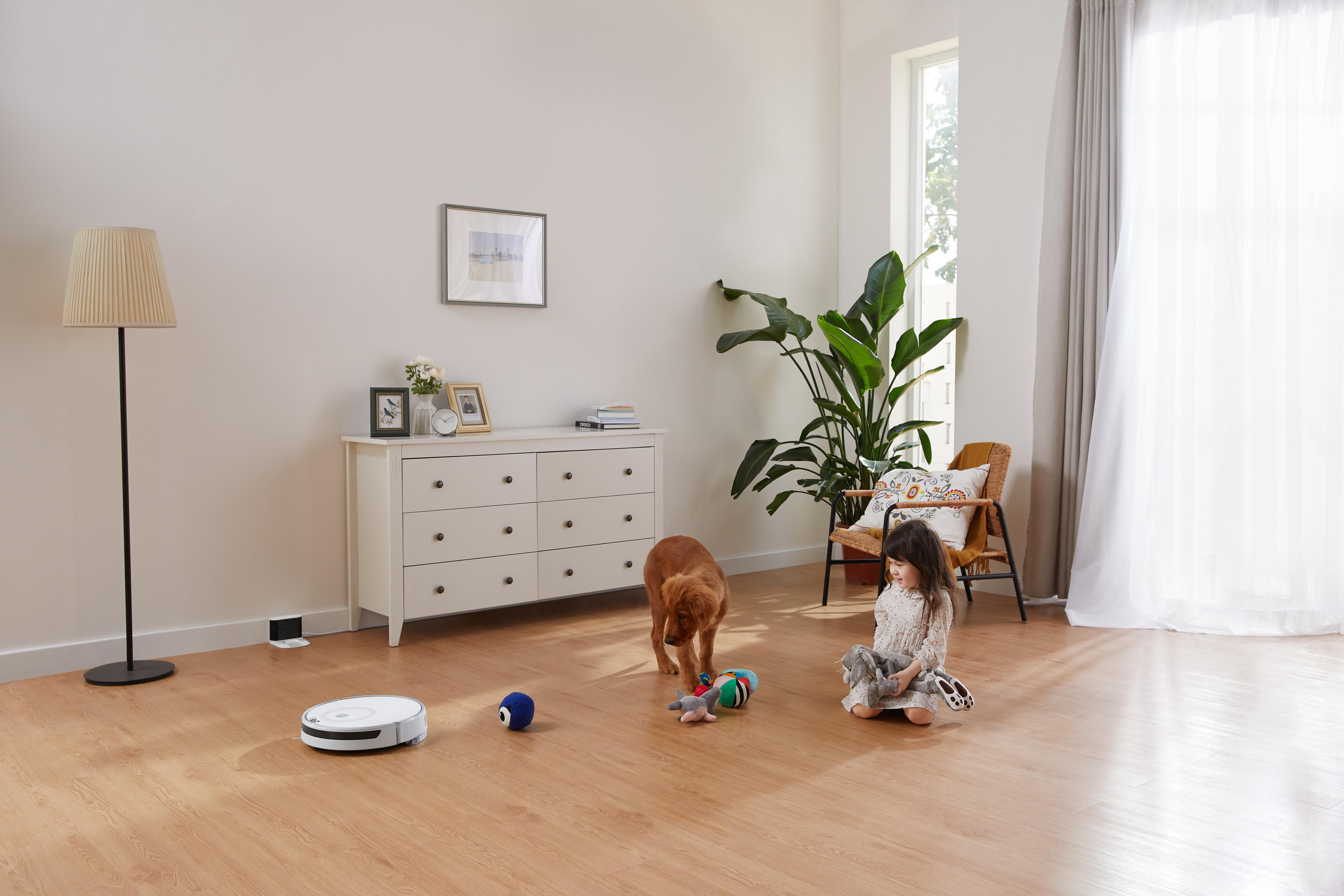 Ein Wohnzimmer mit einem Mädchen und einem Hund. Ein Roboterstaubsauger steht auf dem Boden.