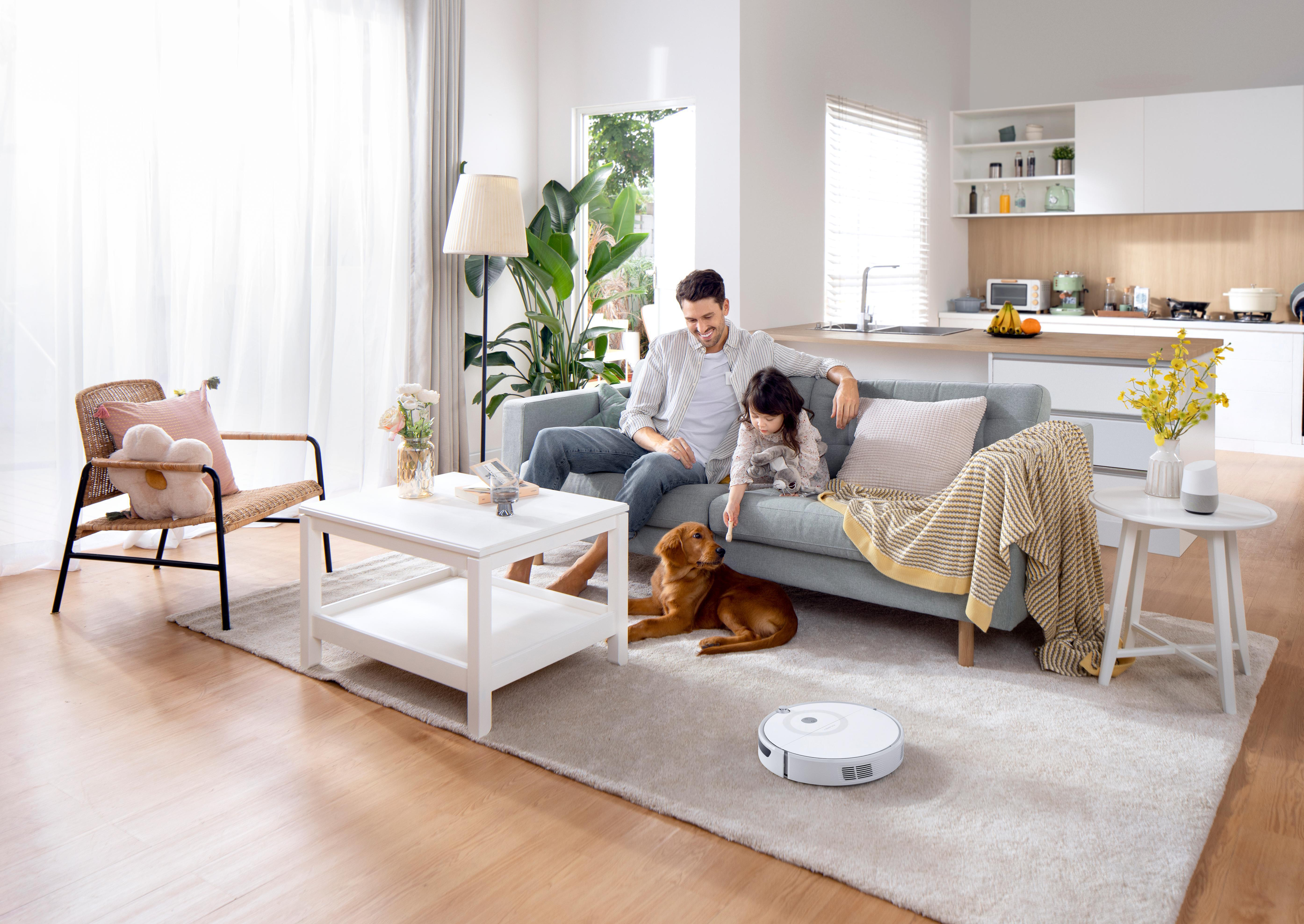 Eine Familie sitzt mit einem Hund auf einem Sofa. Ein Roboter-Staubsauger steht auf dem Boden.