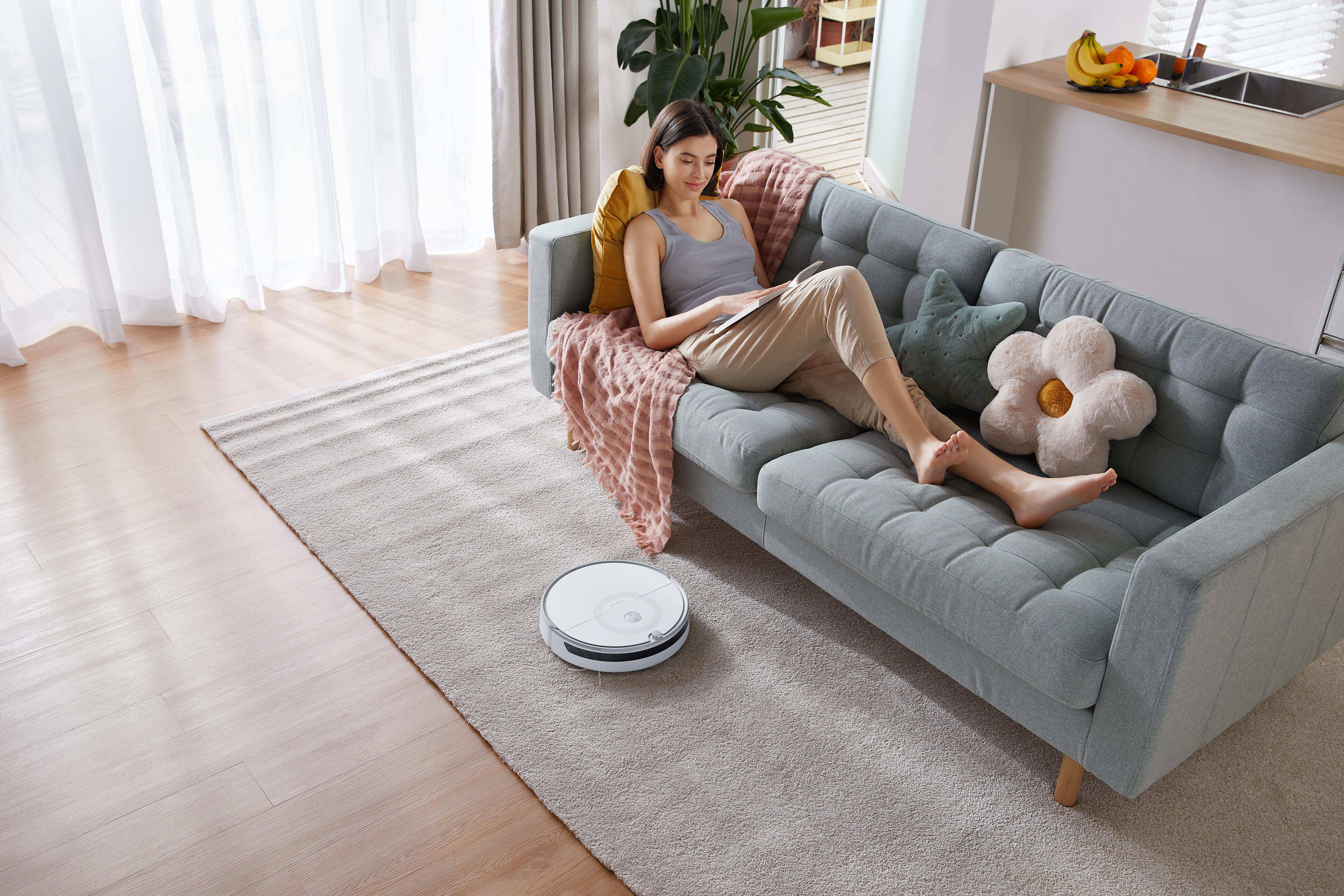 Frau auf dem Sofa mit einem Roboter-Staubsauger auf dem Boden.