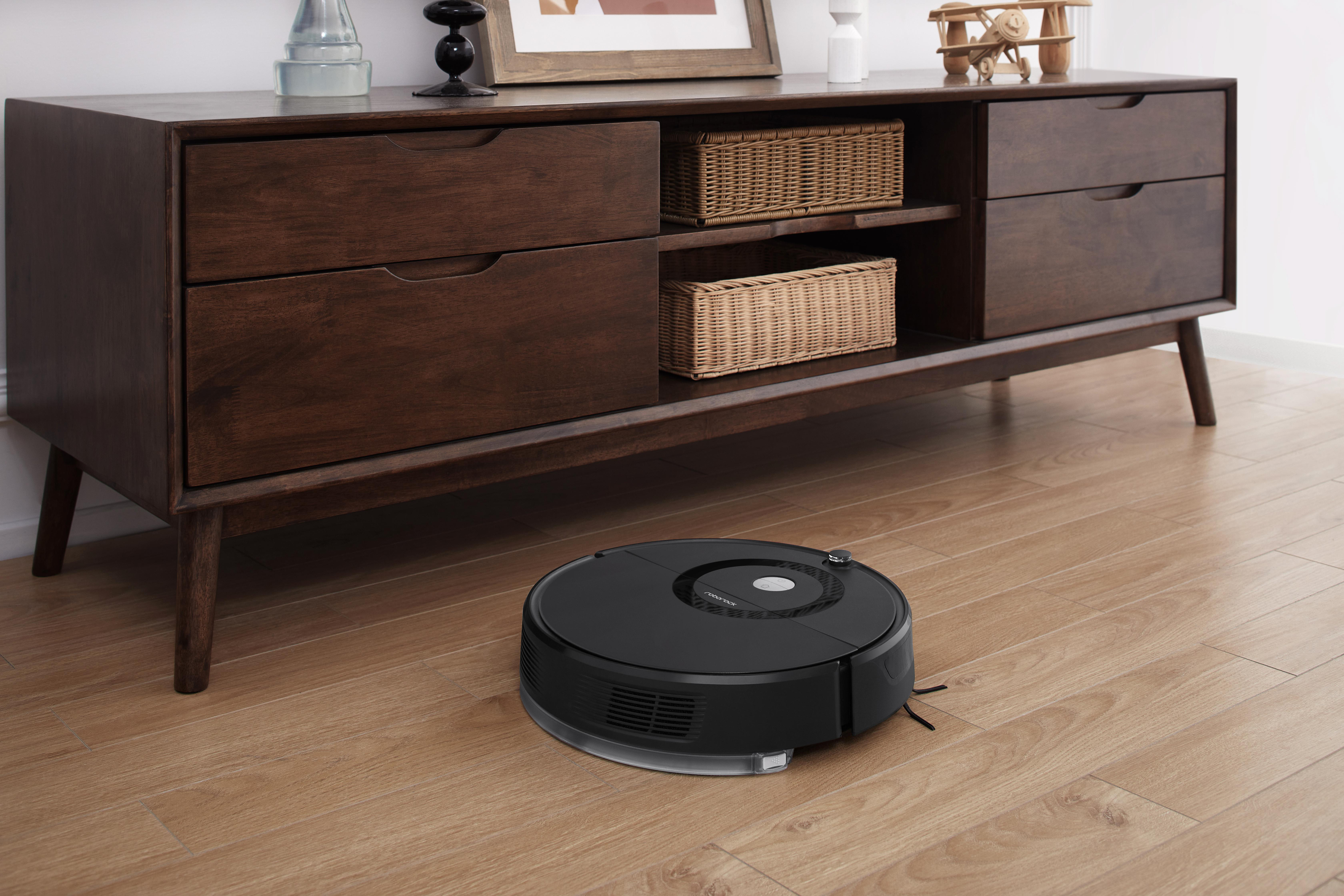 Ein schwarzer Roboter-Staubsauger auf Holzboden vor einem Holzschrank.