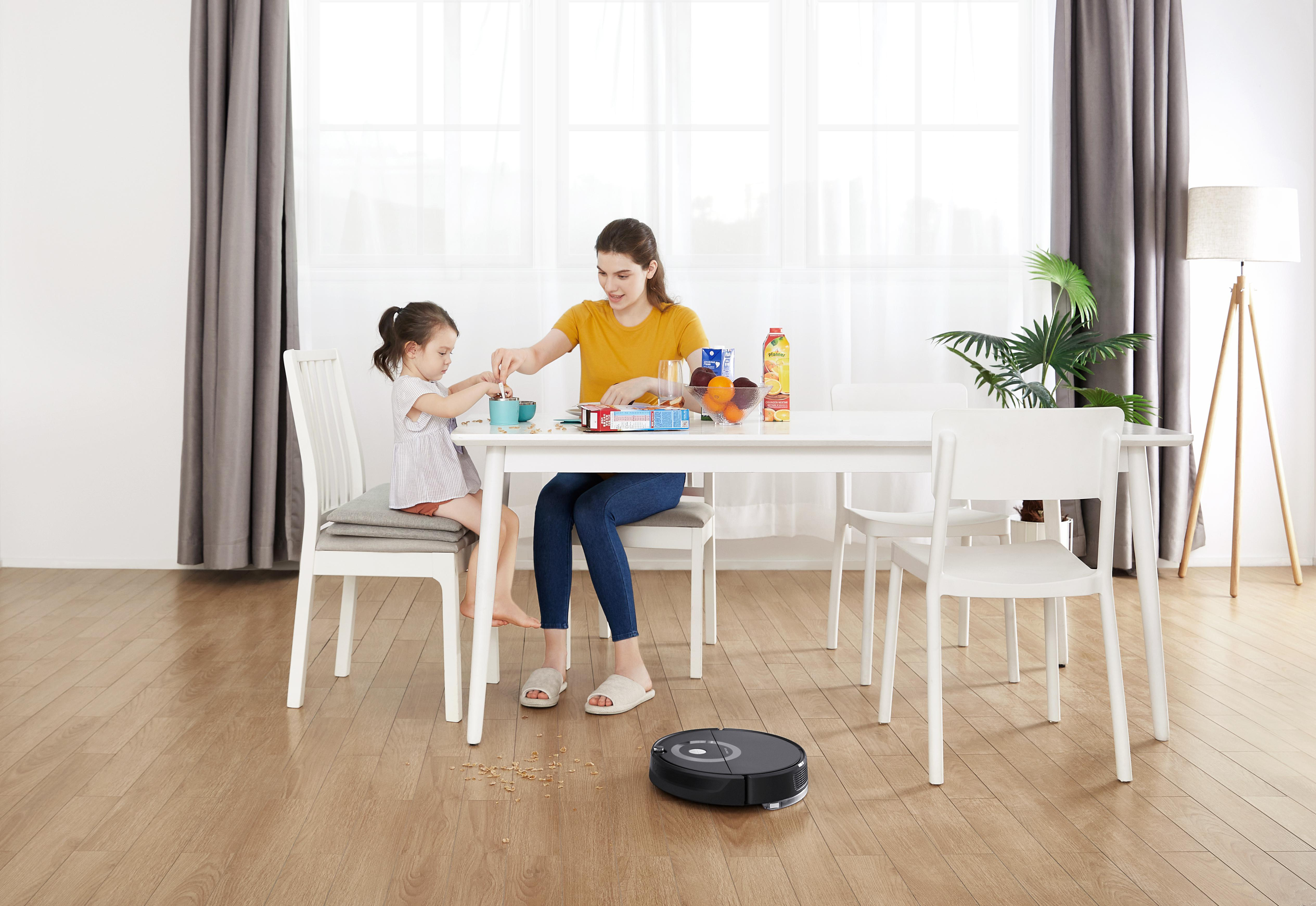 Eine Frau und ein Kind essen an einem Tisch. Ein Roboter-Staubsauger steht auf dem Boden.