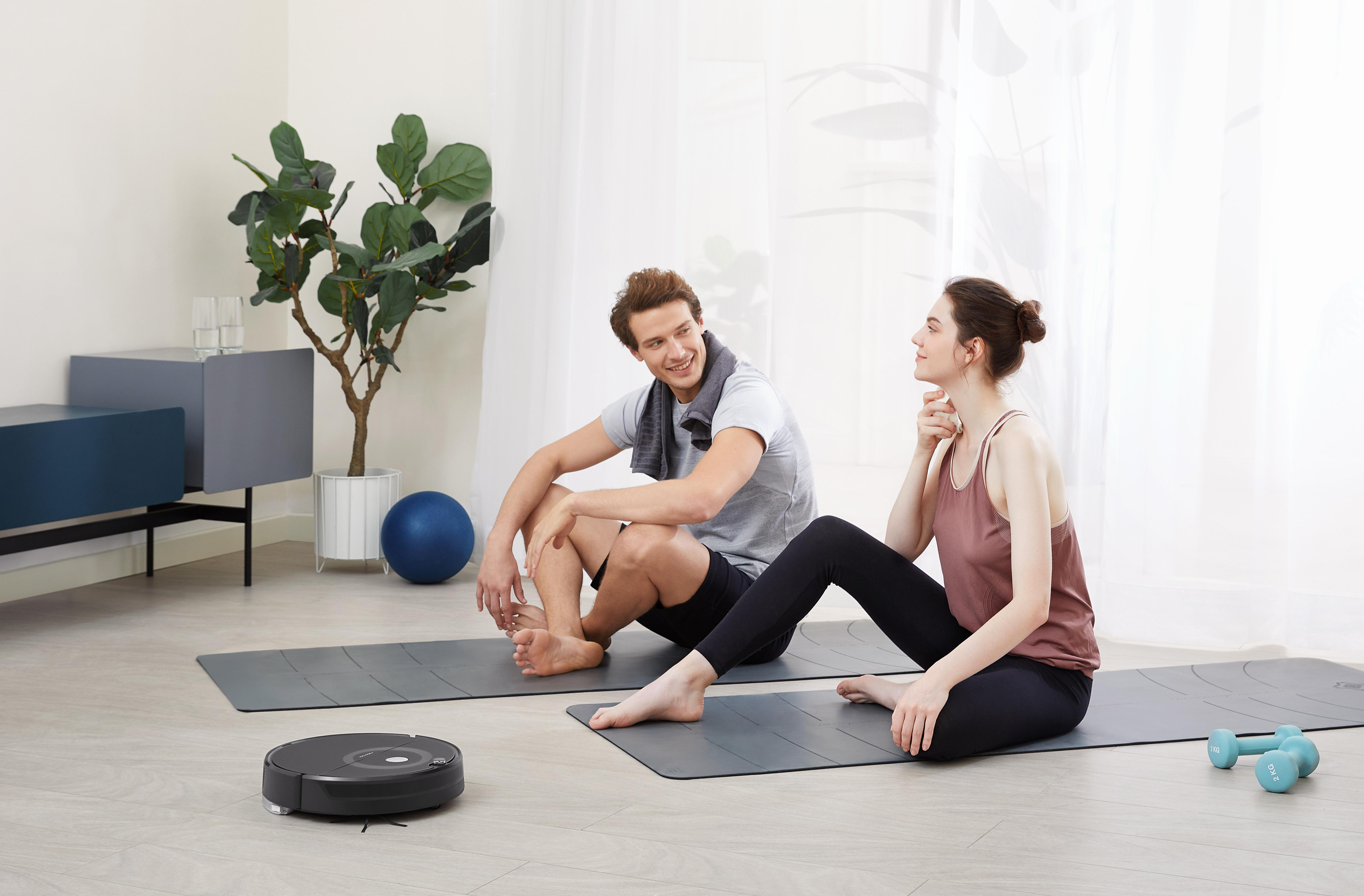 Mann und Frau sitzen auf Matten, in der Nähe eines Staubsaugers. Hintergrund: Fenster, Pflanzen und ein blauer Ball.
