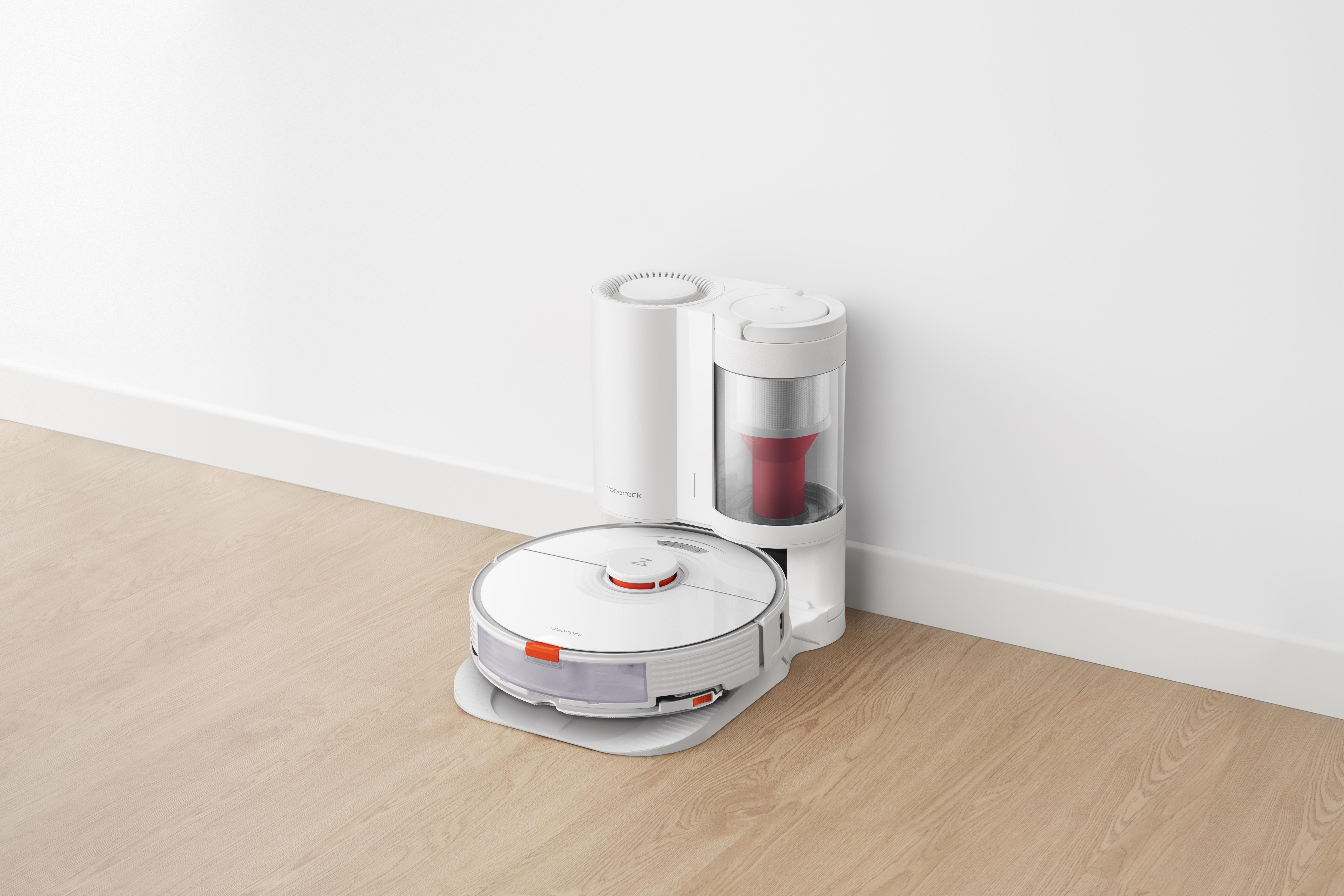 Ein weißer Roboter-Staubsauger und seine Ladestation auf einem Holzboden vor einer weißen Wand.