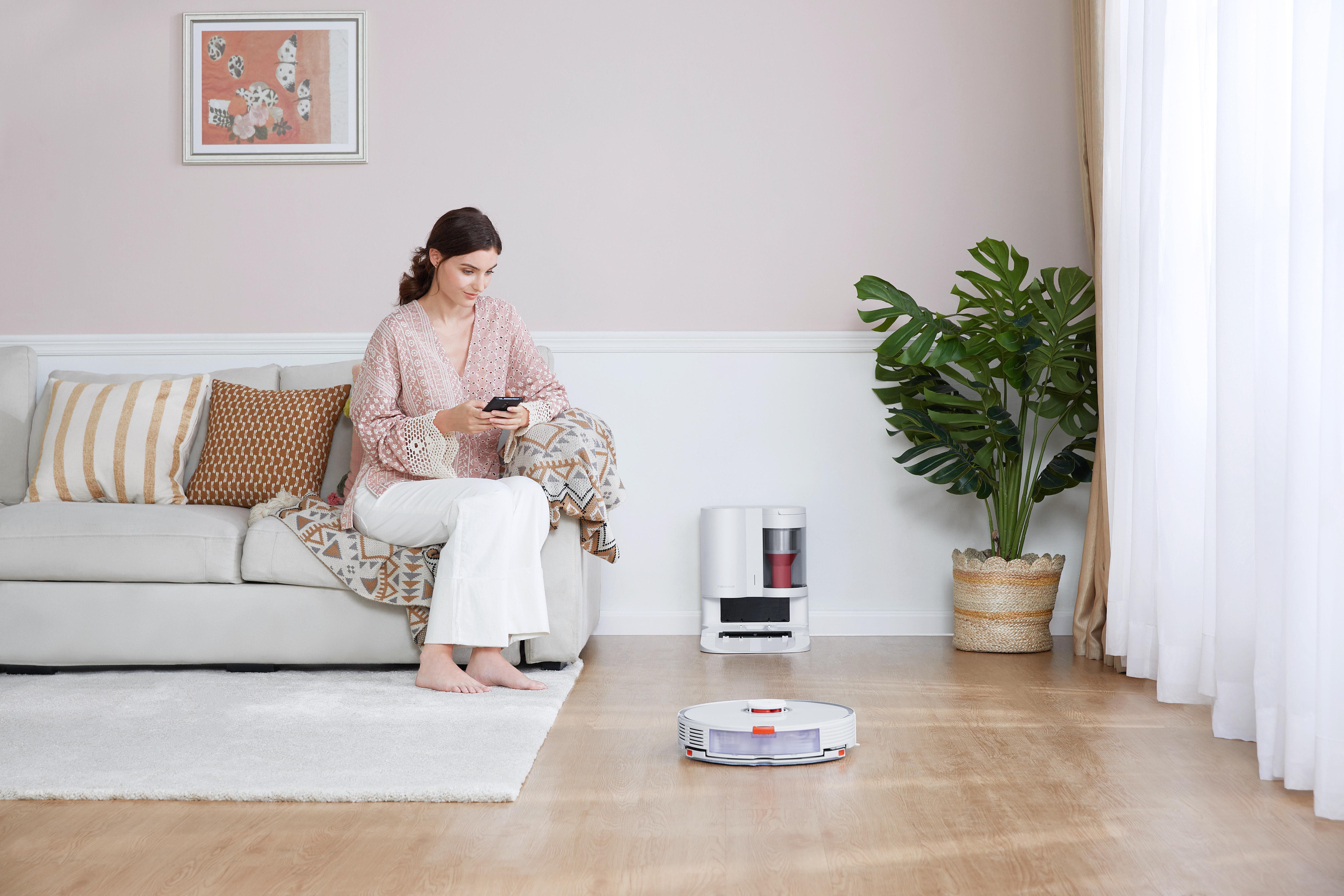 Frau auf einem Sofa, die ein Telefon benutzt, während ein Roboterstaubsauger den Boden reinigt.