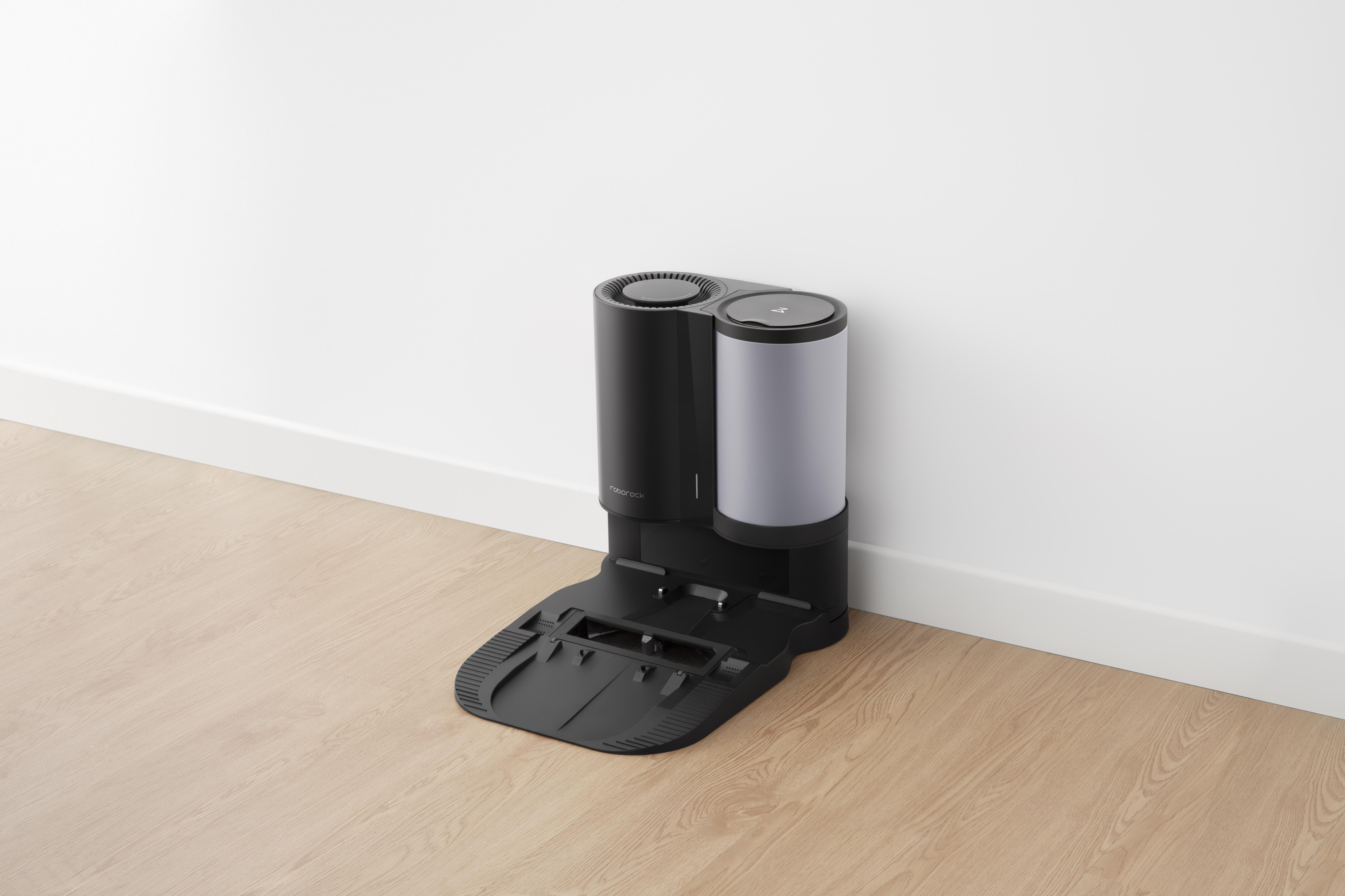 Ein schwarz-grauer Roboter-Staubsauger und eine Ladestation vor einer weißen Wand.