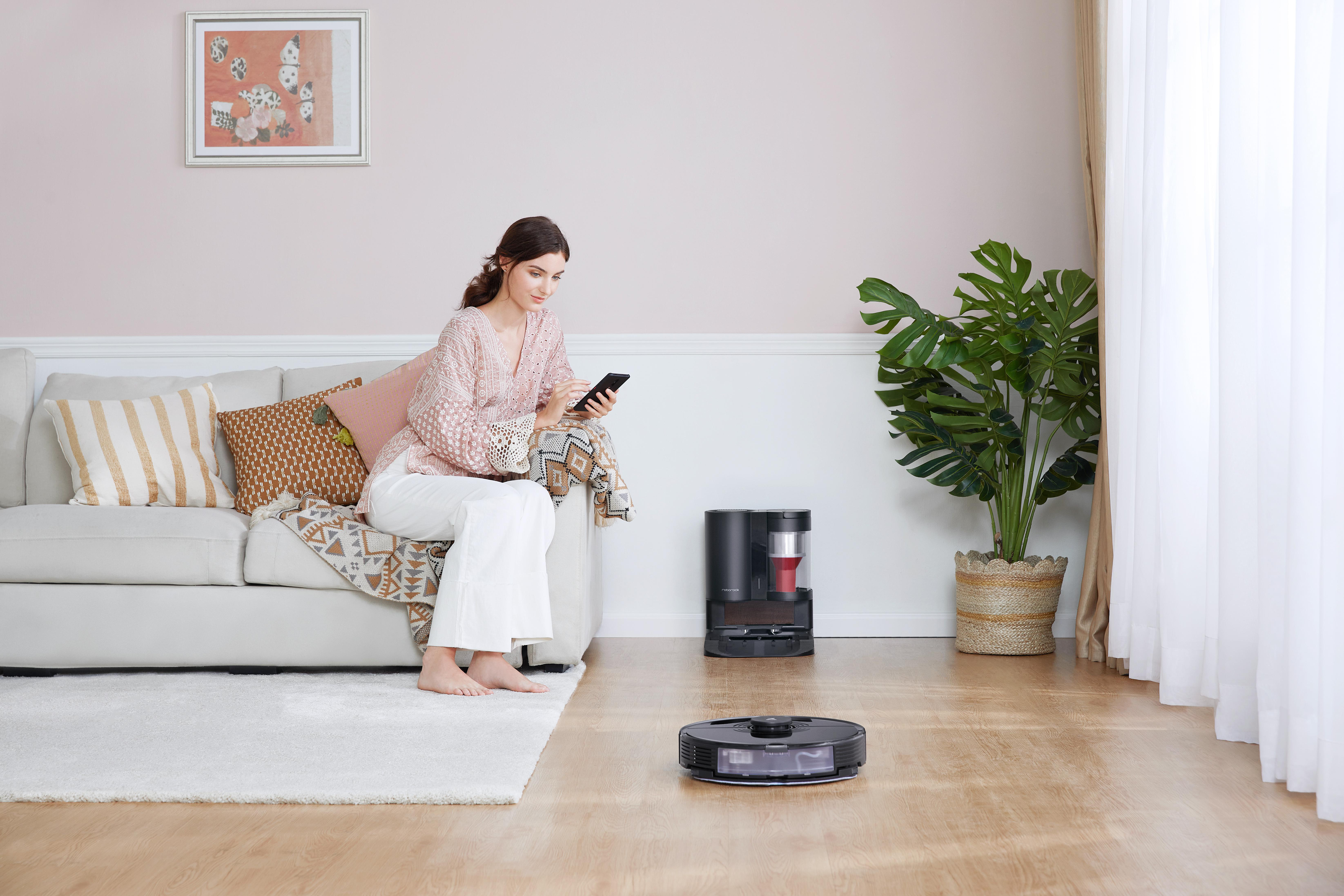 Frau benutzt Telefon auf Sofa, während Roboter-Staubsauger auf dem Boden arbeitet.