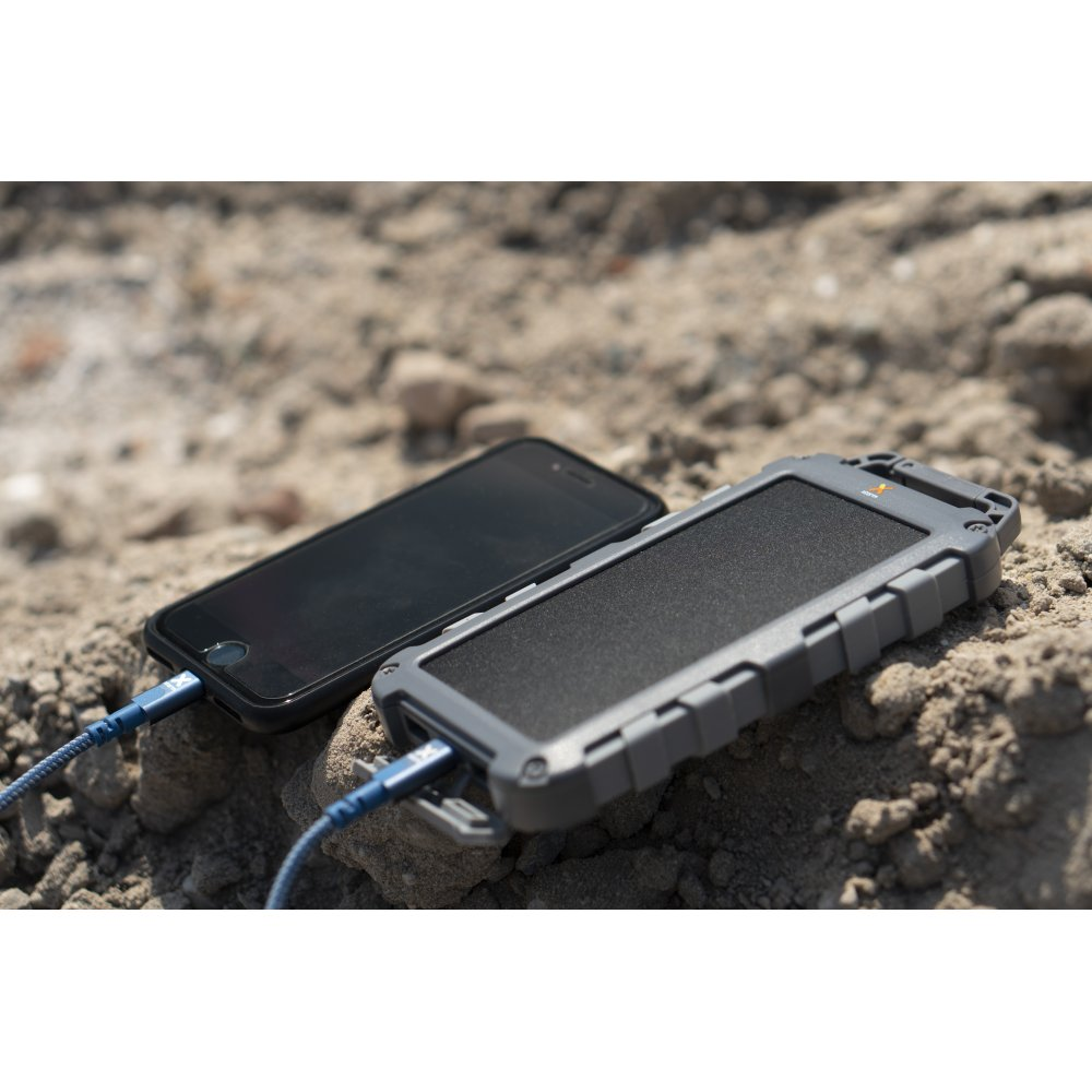Ein schwarzes Smartphone, verbunden mit einer grauen Powerbank mit blauem Kabel, auf rauer, steinbedeckter Oberfläche.