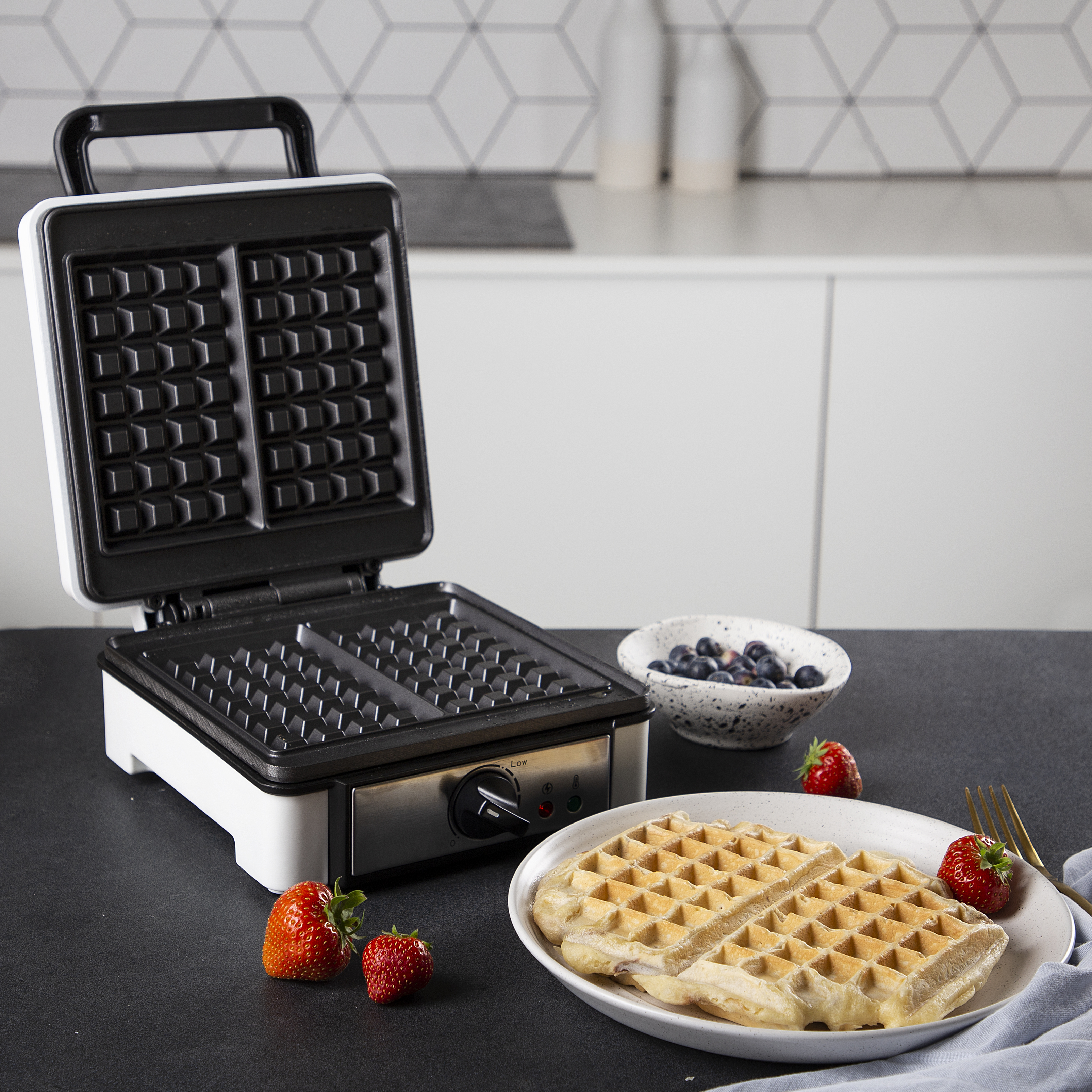 Wafelijzer met wafels. Bord met wafels, kom met blauwe bessen, aardbeien, op een zwart oppervlak.