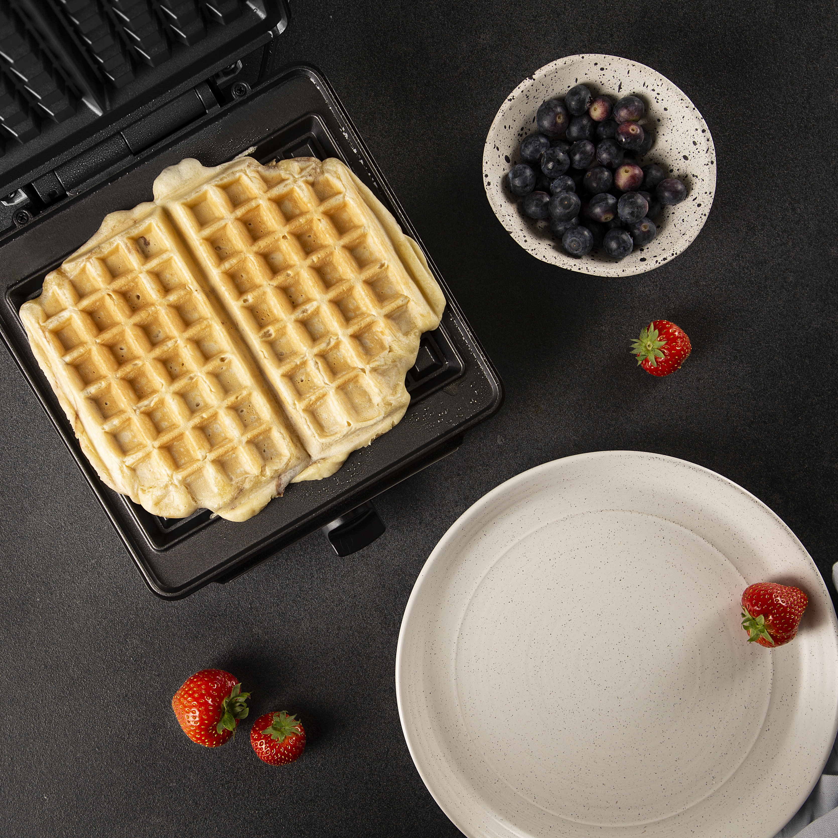 Wafels in een wafelijzer, een kom met blauwe bessen, aardbeien en een bord.