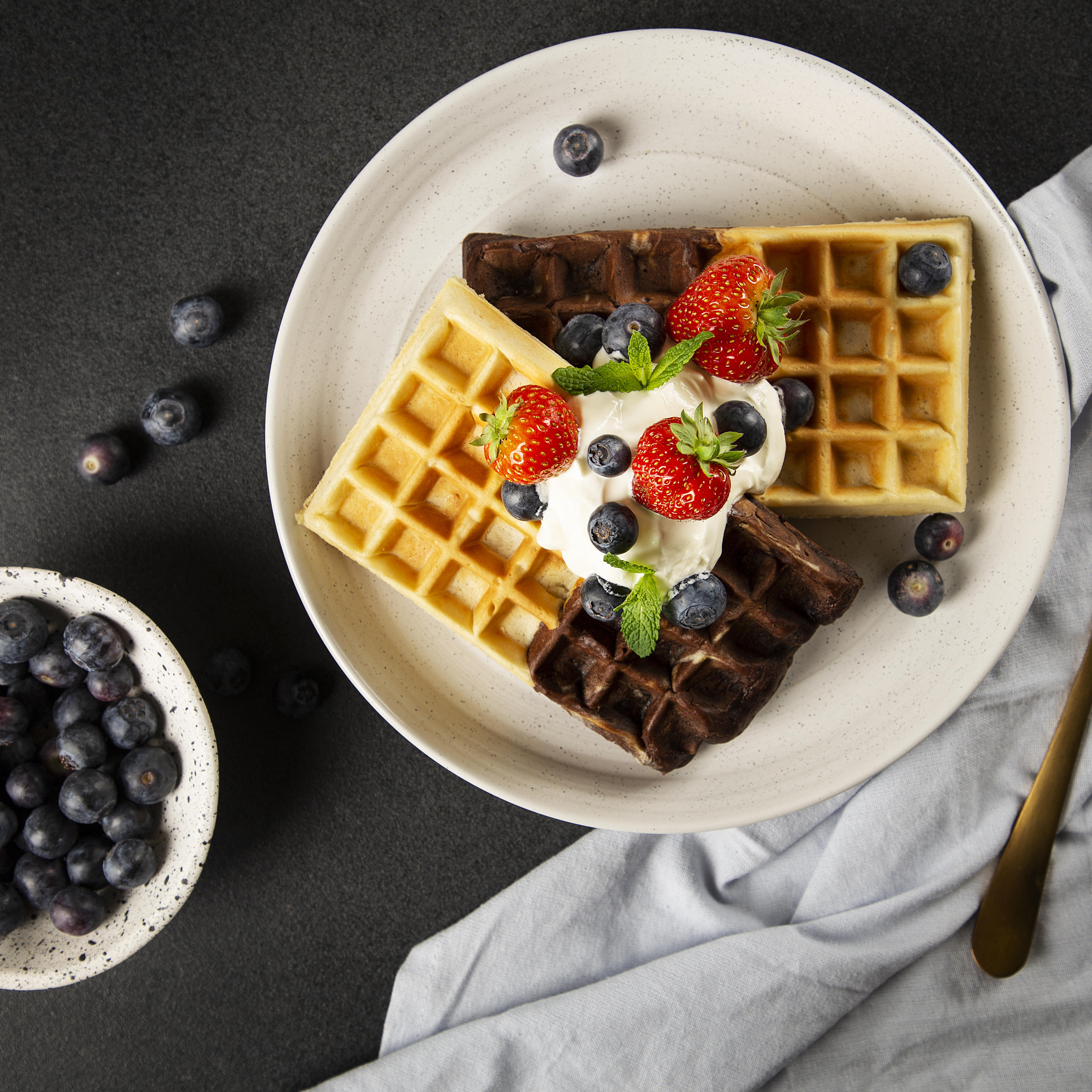 Wafels met bosbessen, aardbeien en room op een bord, met een kom bosbessen. Zwarte achtergrond.