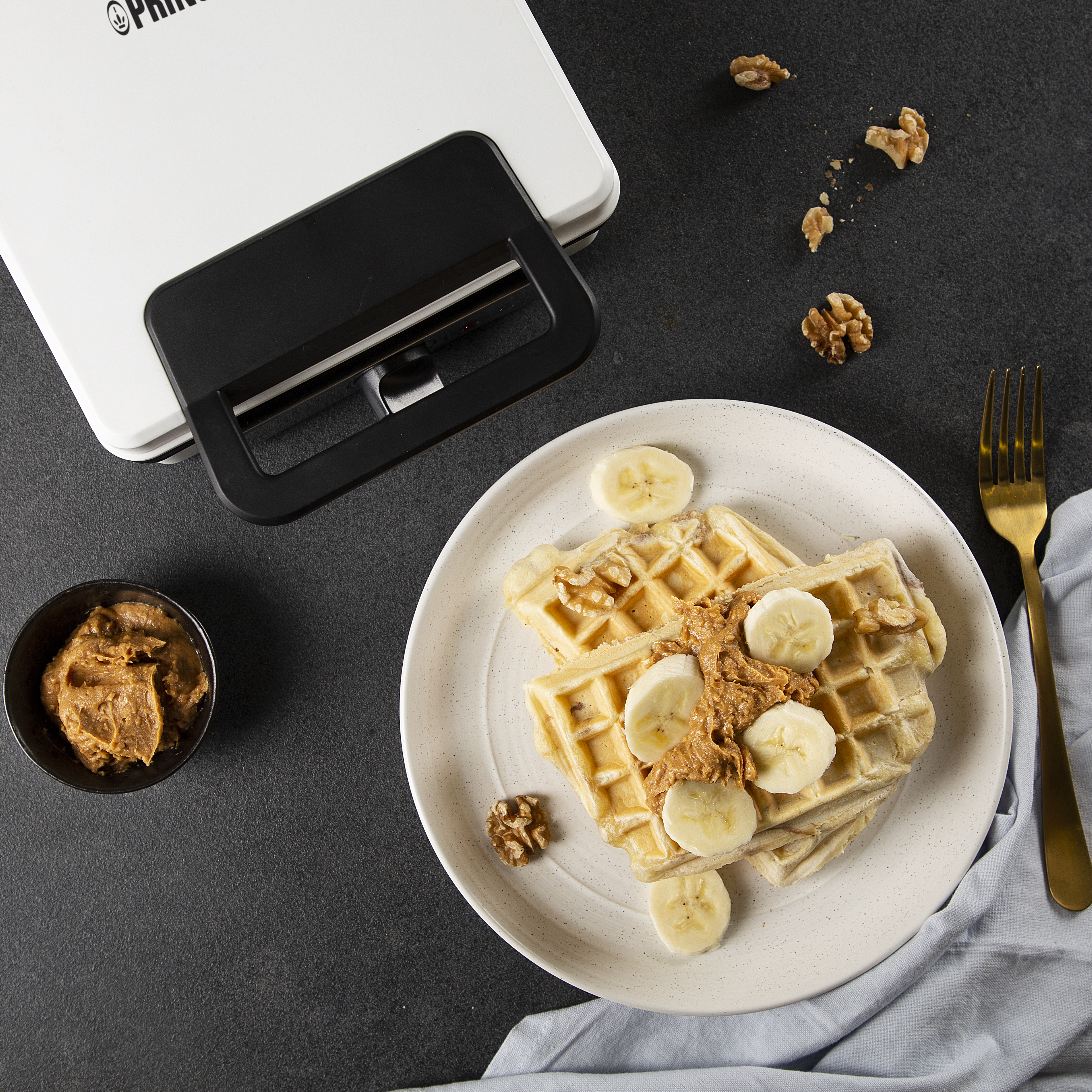 Wafels met plakjes banaan, pindakaas en walnoten op een bord, met een wafelijzer op de achtergrond.