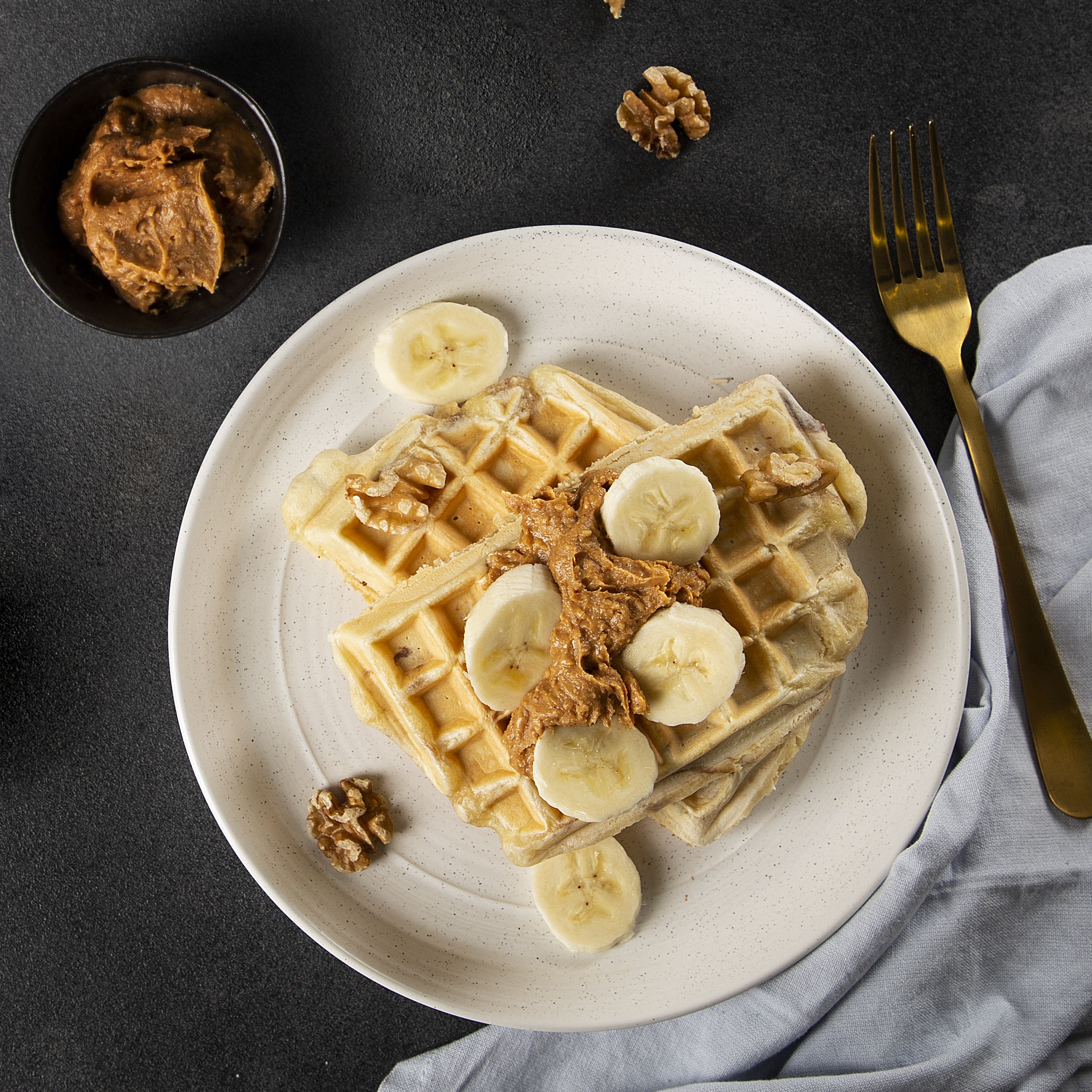 Wafels met bananenschijfjes en notenboter op een bord, met walnoten en een vork.