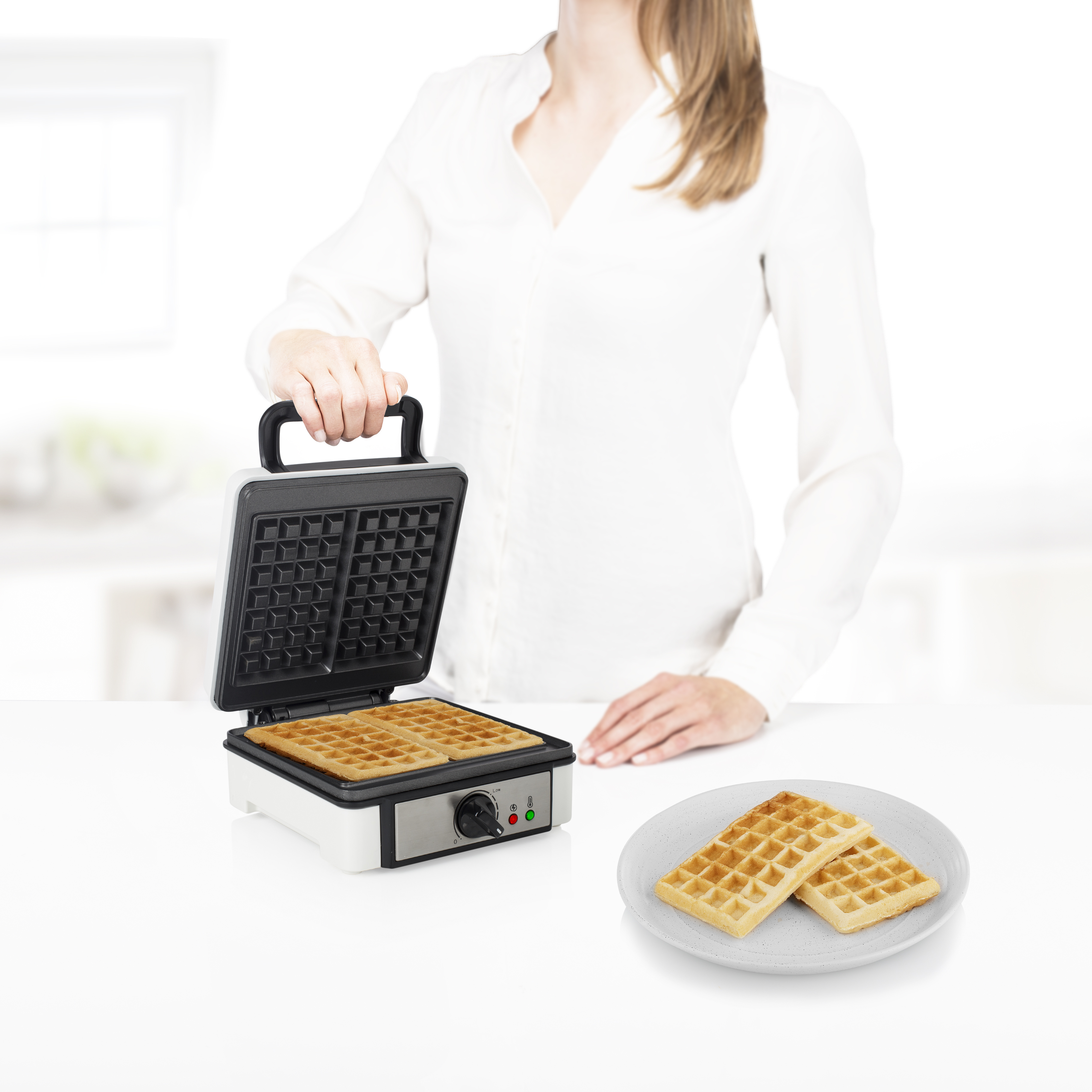 Een vrouw houdt een wafelijzer vast met wafels erin en twee wafels op een bord.