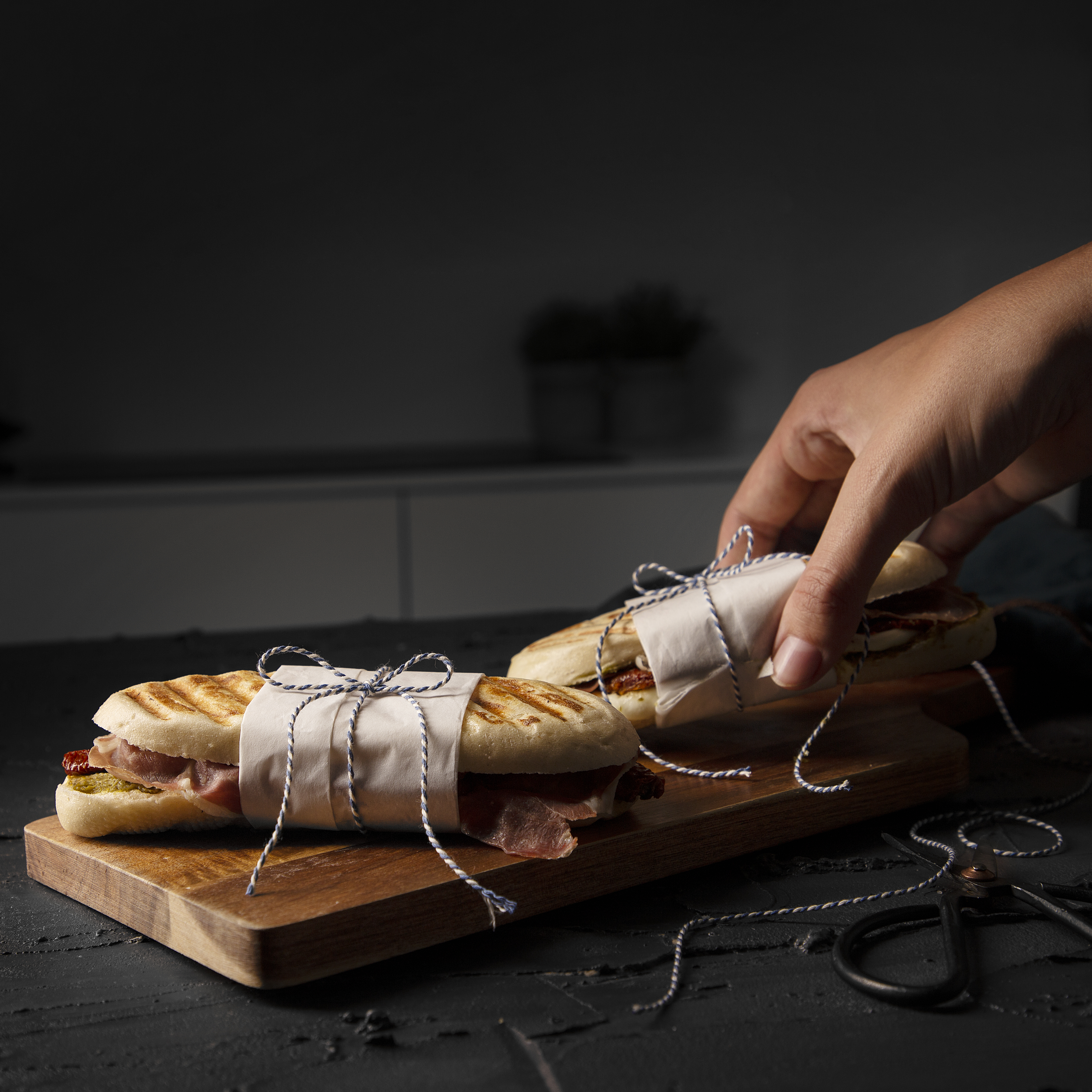 Two sandwiches wrapped in paper, tied with twine, on a wooden board. A hand reaches for one.