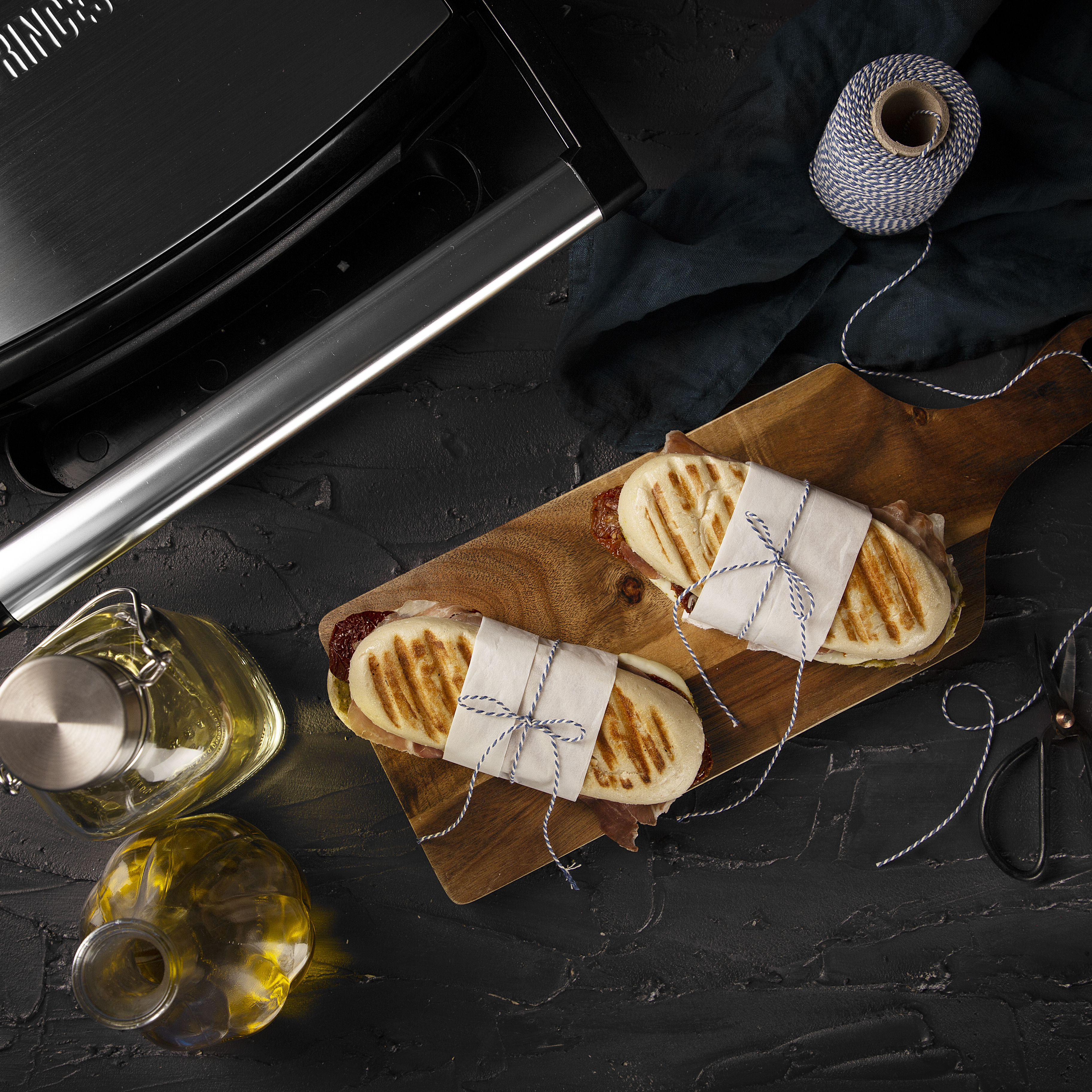 Panini sandwiches on a wooden board, with oil, string, and a panini press in the background.