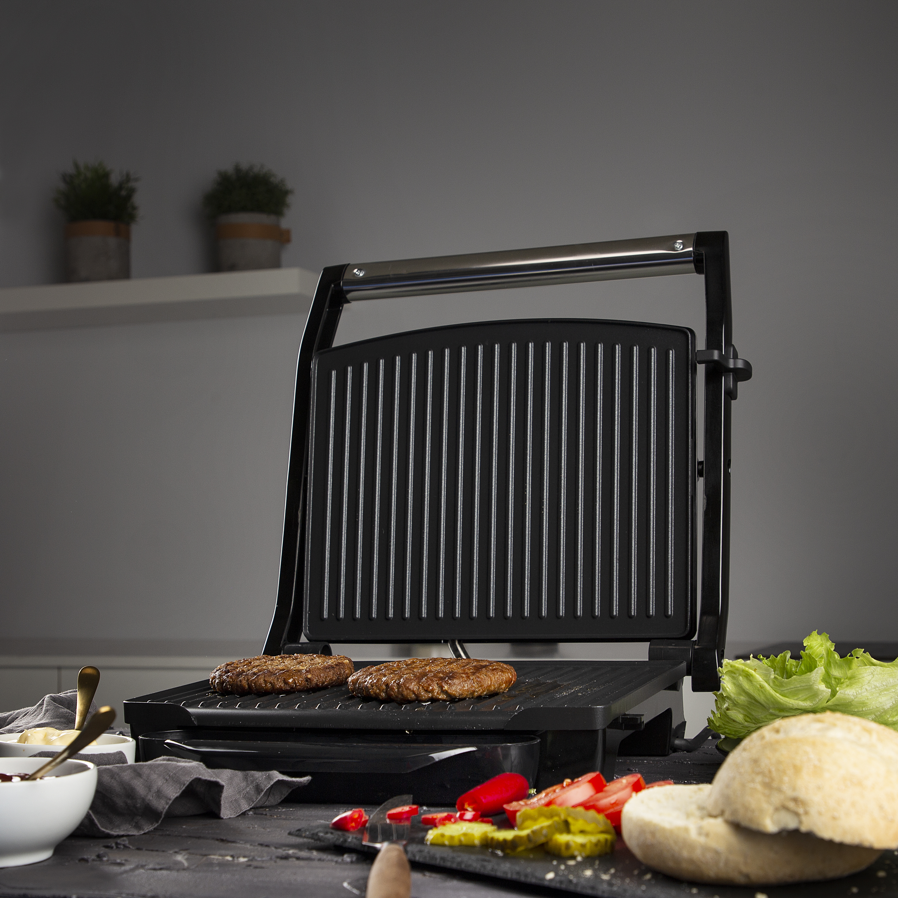 Grill with two burgers cooking with sides of salad, tomatoes, and bread.