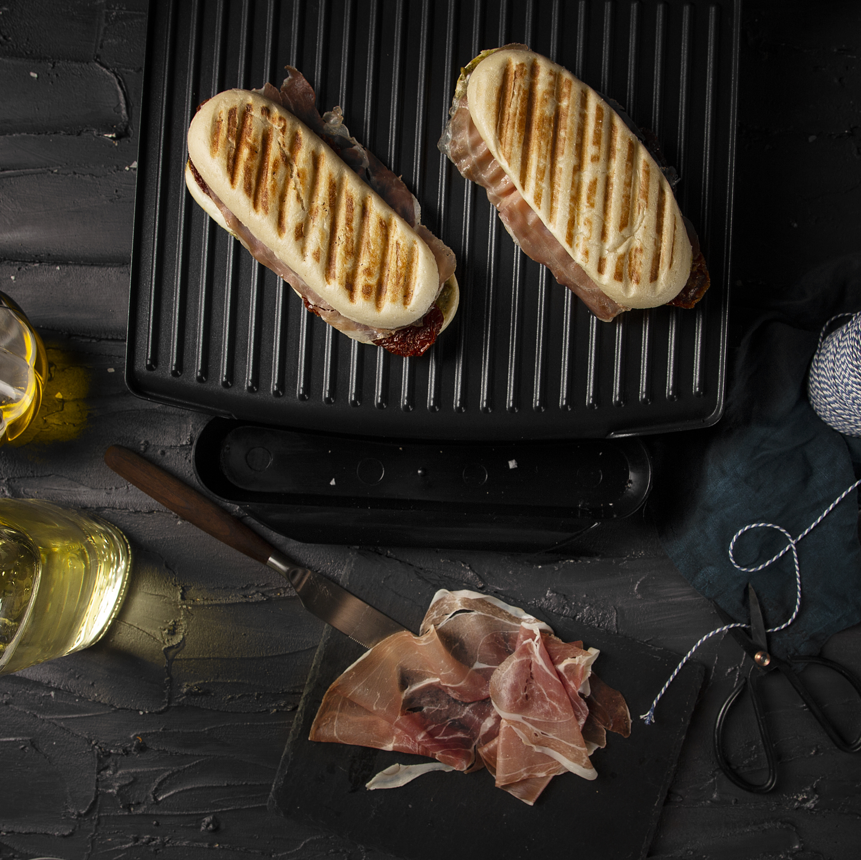 Two grilled sandwiches on a grill next to ingredients, including prosciutto and oil.