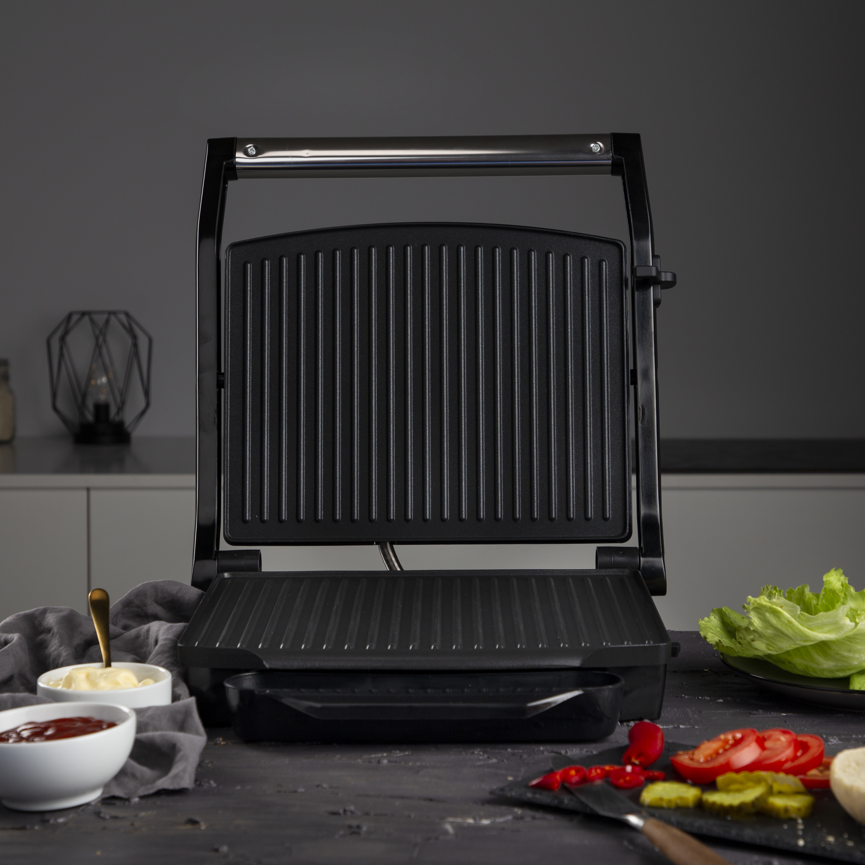 A black sandwich maker open with a tray of food in front.