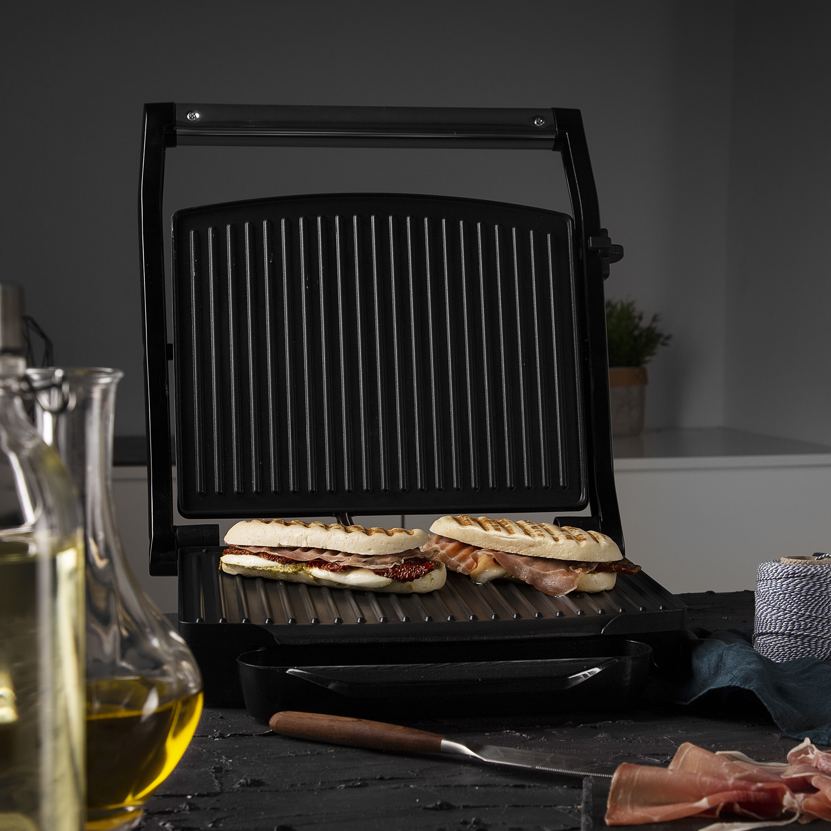 Panini grill with two sandwiches, olive oil, and a knife on a dark surface.