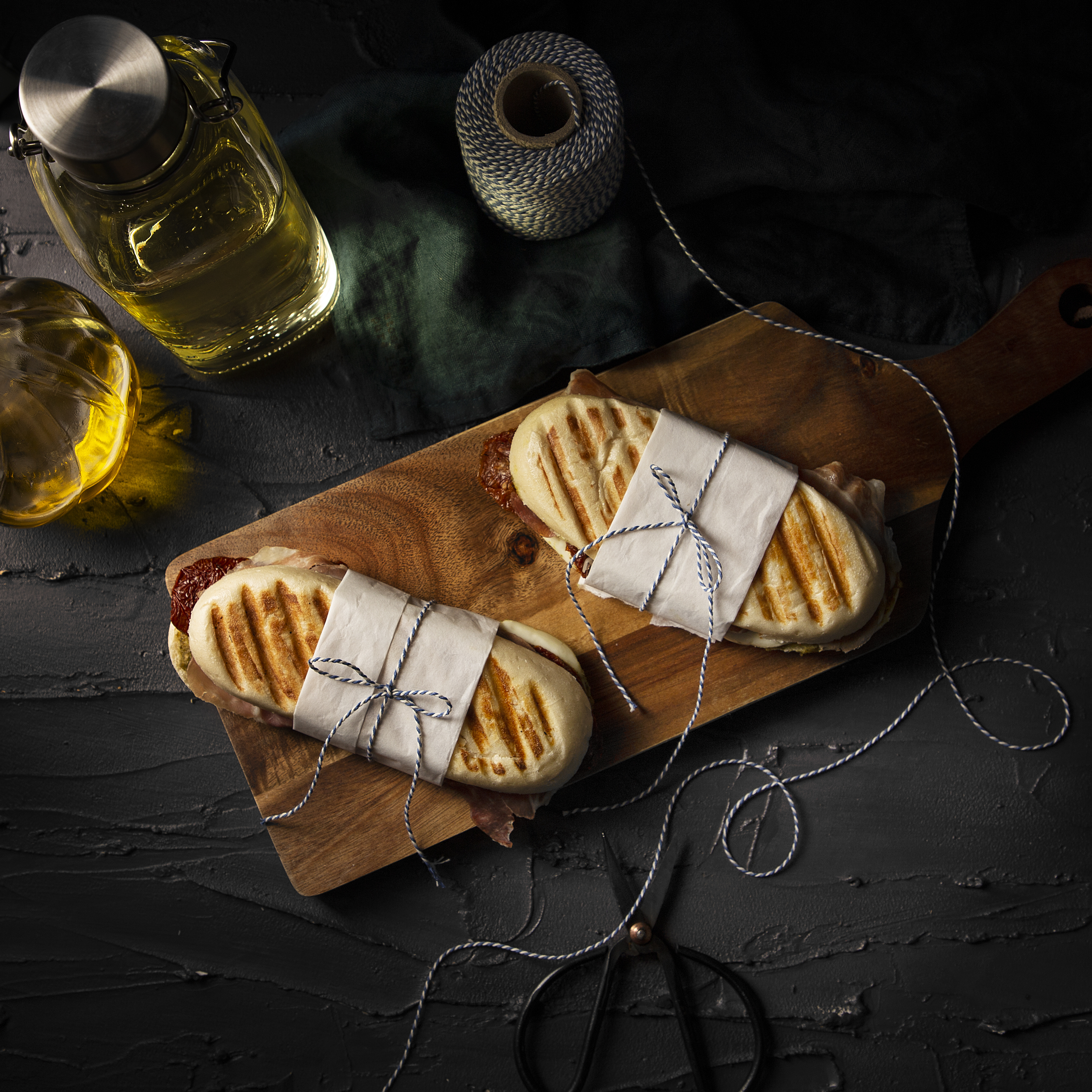 Two panini sandwiches wrapped in paper and tied with string on a wooden board.