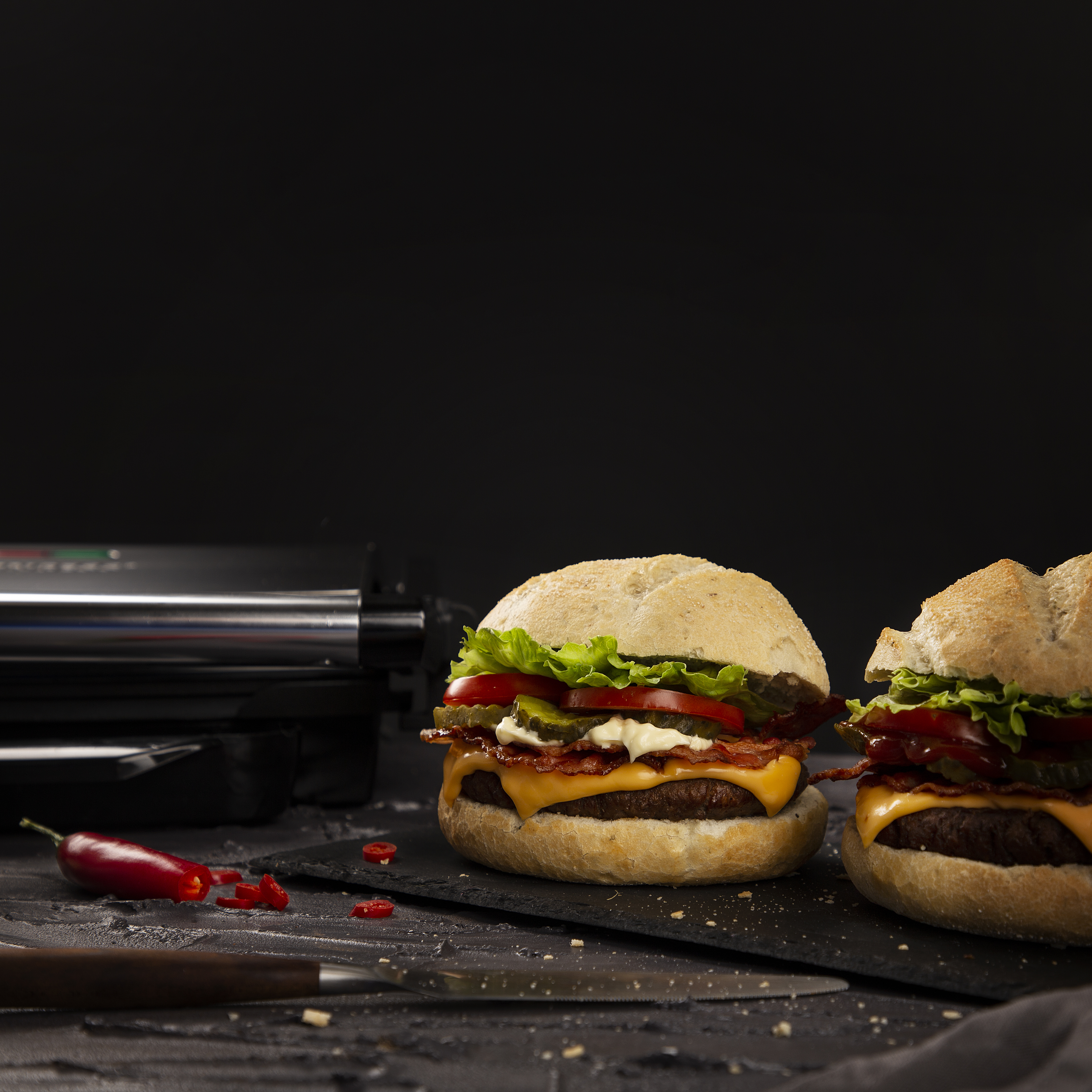 Two burgers on a black board with a grill in the background. Red pepper and knife.