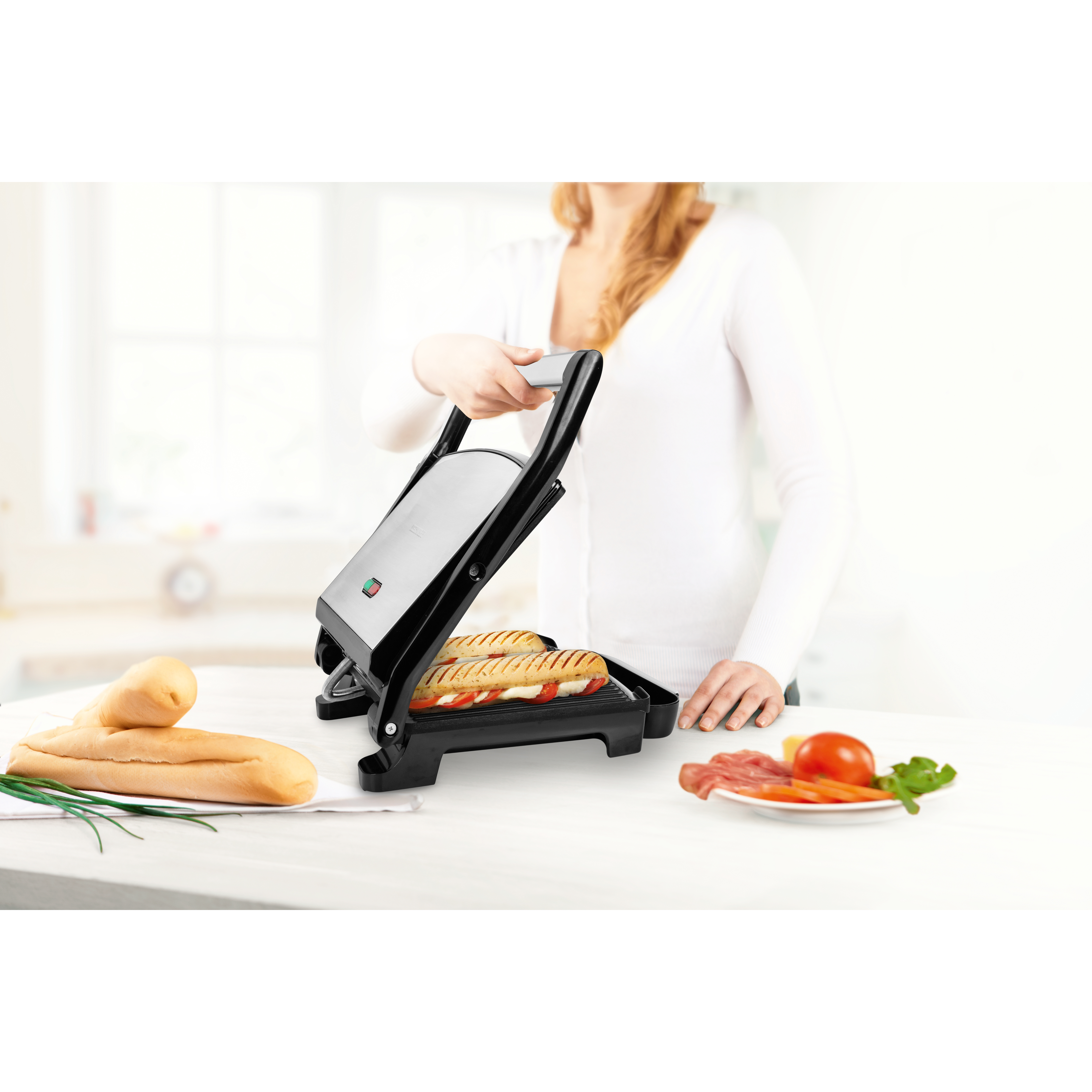 A person using a sandwich maker with sandwiches and ingredients on a table.