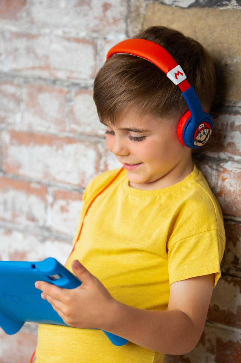 Un ragazzino con cuffie di Mario guarda un tablet blu. Appoggiato a un muro di mattoni.