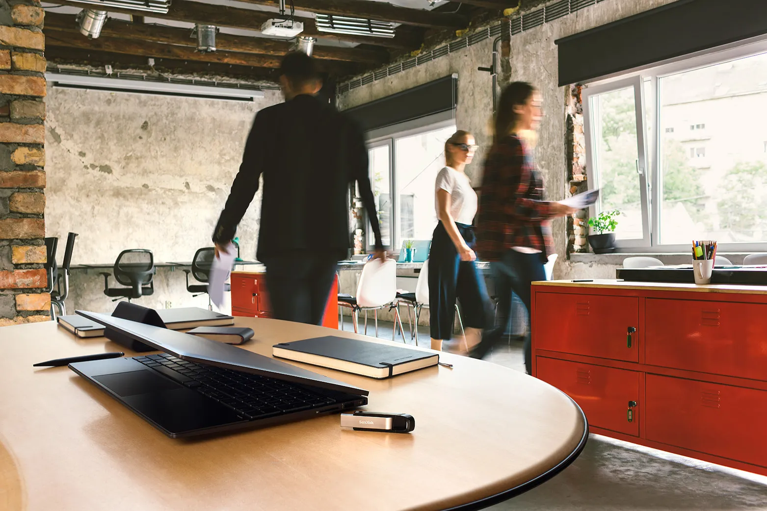 Un ordinateur portable sur une table en bois dans un bureau, avec des gens qui marchent en arrière-plan.