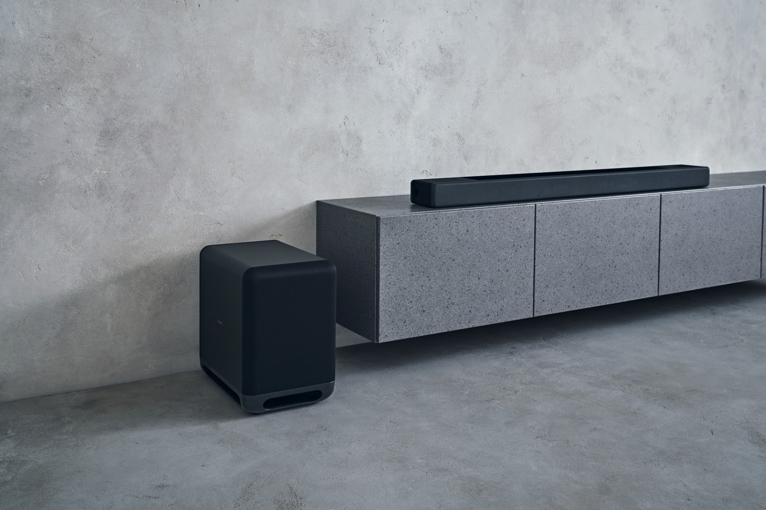 Una configuración de cine en casa con subwoofer, barra de sonido y mueble gris.