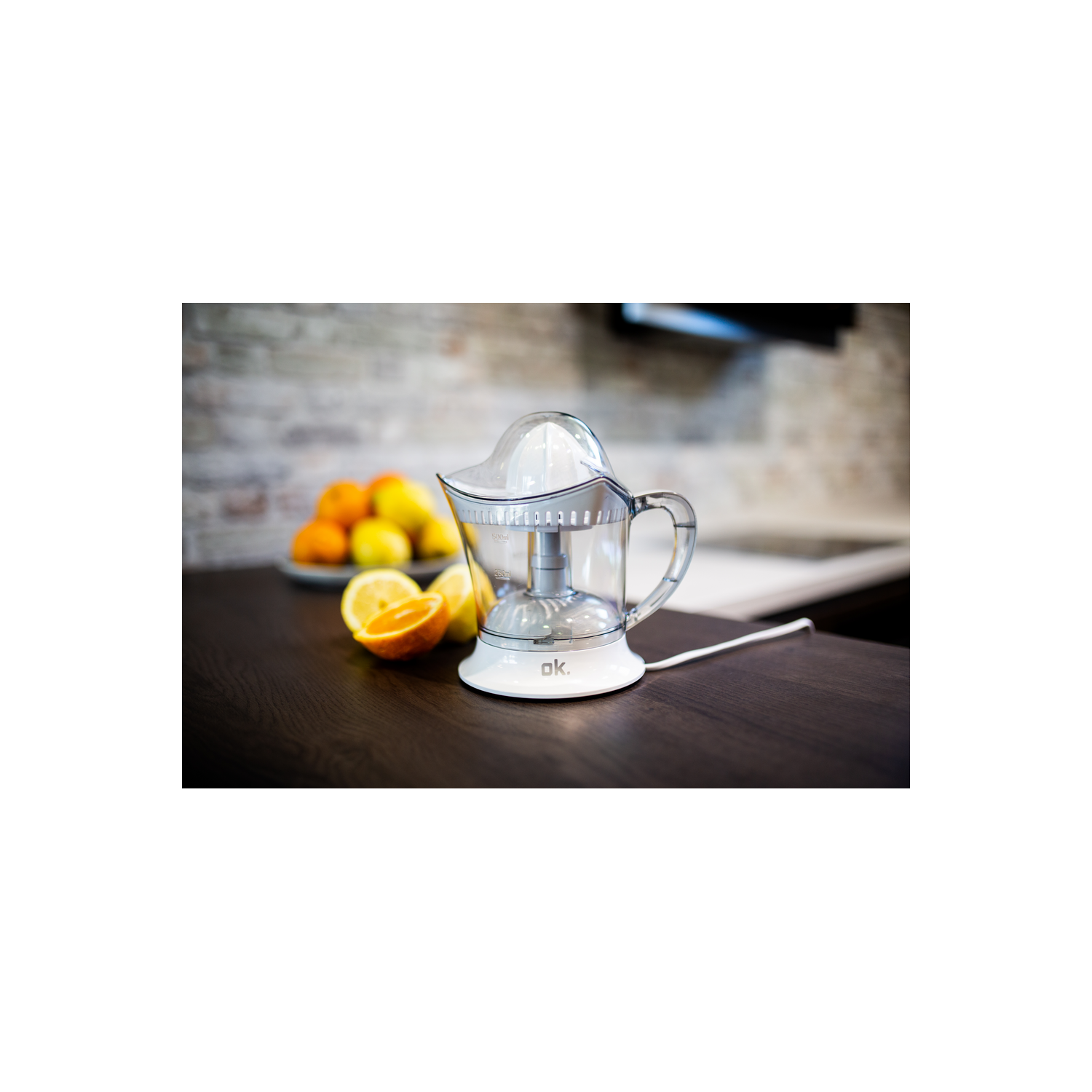 Exprimidor eléctrico con jarra transparente y asa, rodeado de naranjas y un limón.