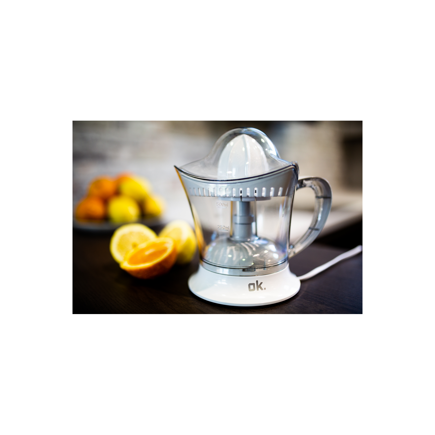 Un exprimidor eléctrico transparente con base blanca, naranjas en rodajas y un plato de naranjas en el fondo.
