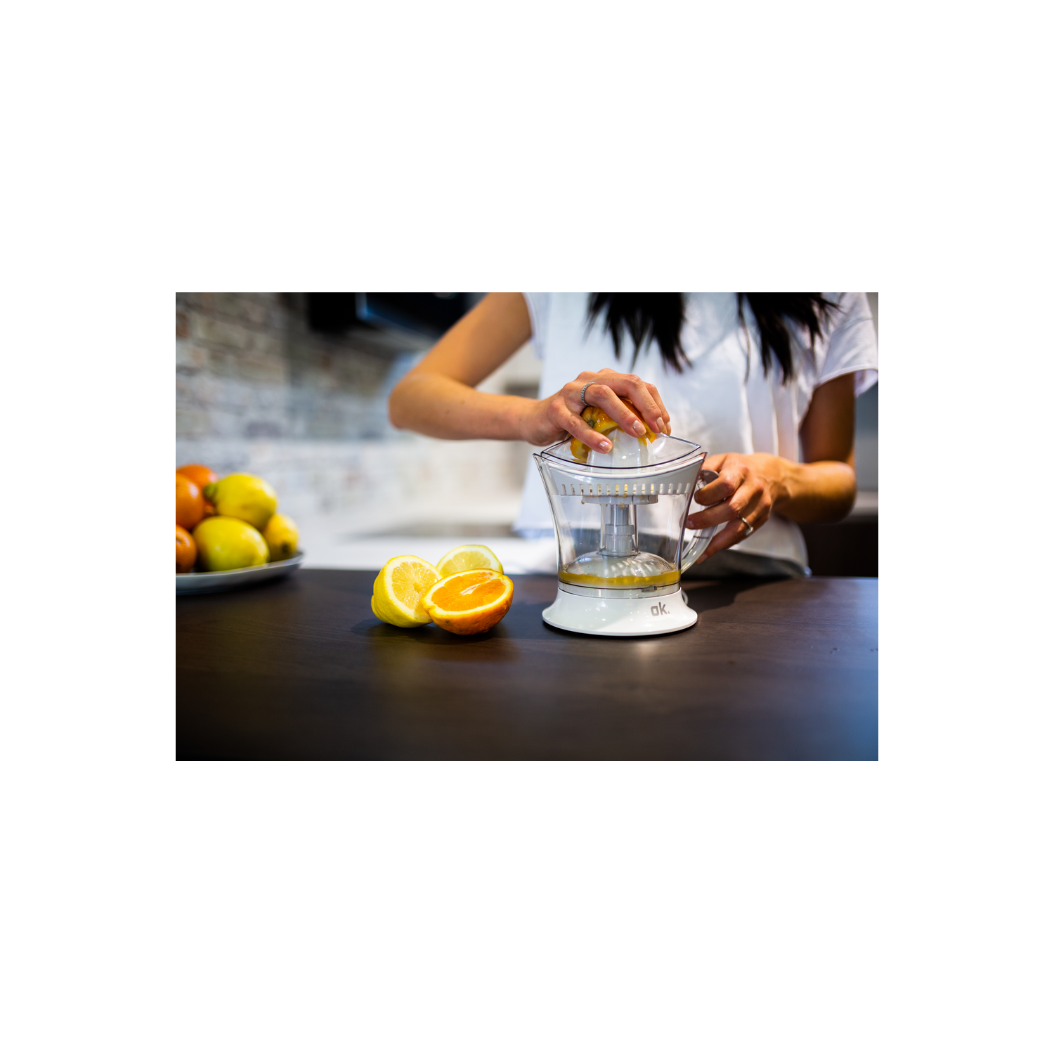 Mujer exprimiendo limones con una exprimidora blanca, con limones y naranjas en una mesa.