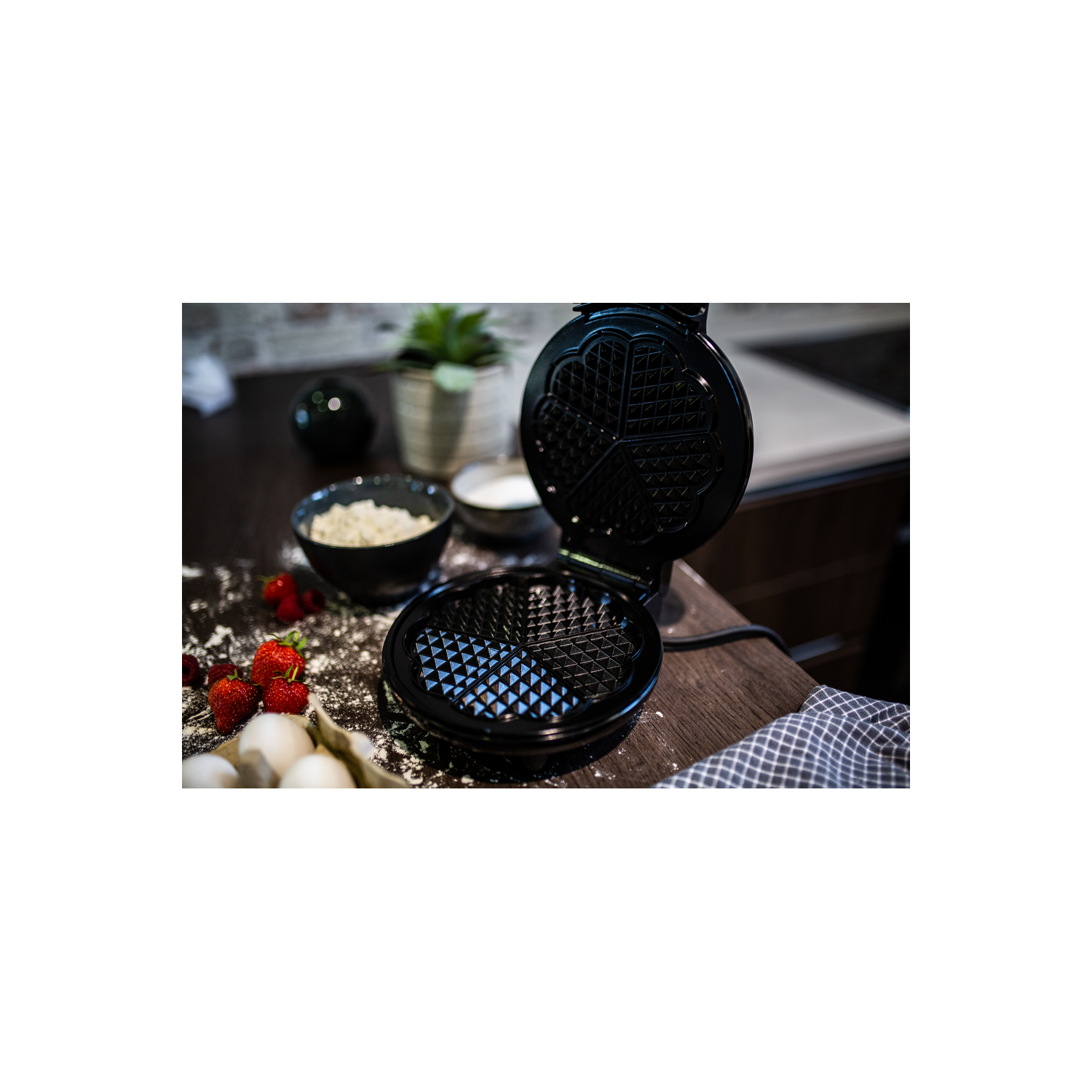 Una gofrera negra con tapa abierta, fresas, huevos y avena sobre una mesa.