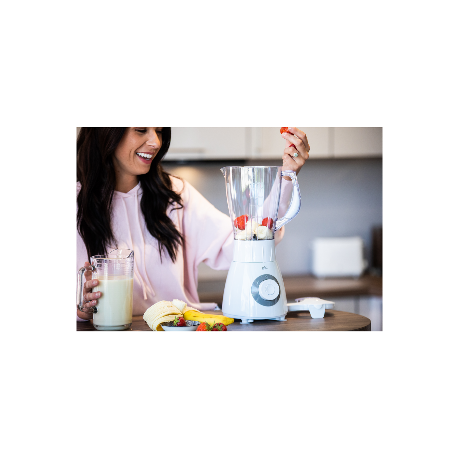 Una mujer preparando un batido con frutas en una licuadora blanca, junto a un vaso de leche.
