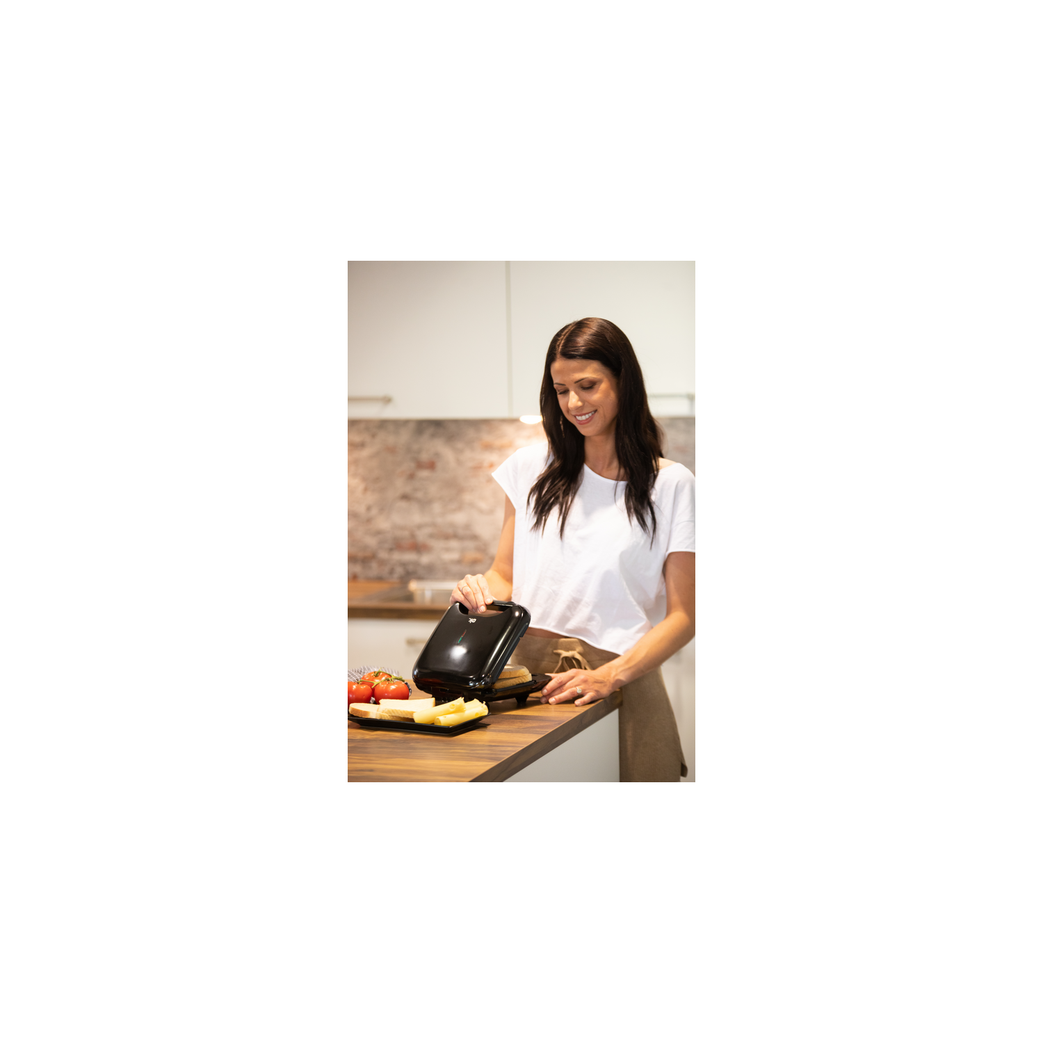 Mujer sosteniendo un procesador de alimentos negro, colocando artículos en una encimera con tomates y queso. Cocina.