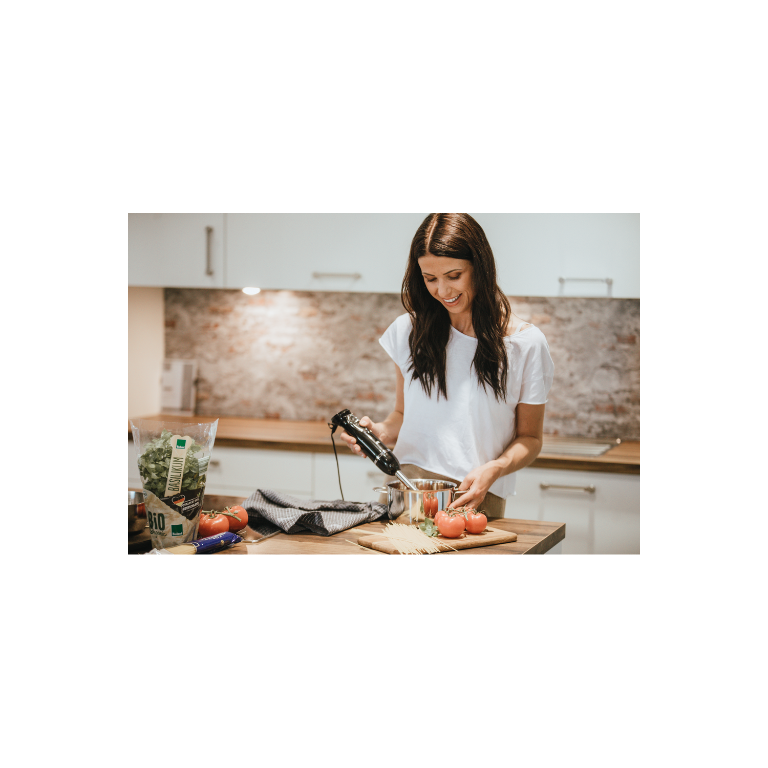 Una mujer con una camisa blanca está cocinando. Está usando una batidora de mano. Se ven tomates y pasta.
