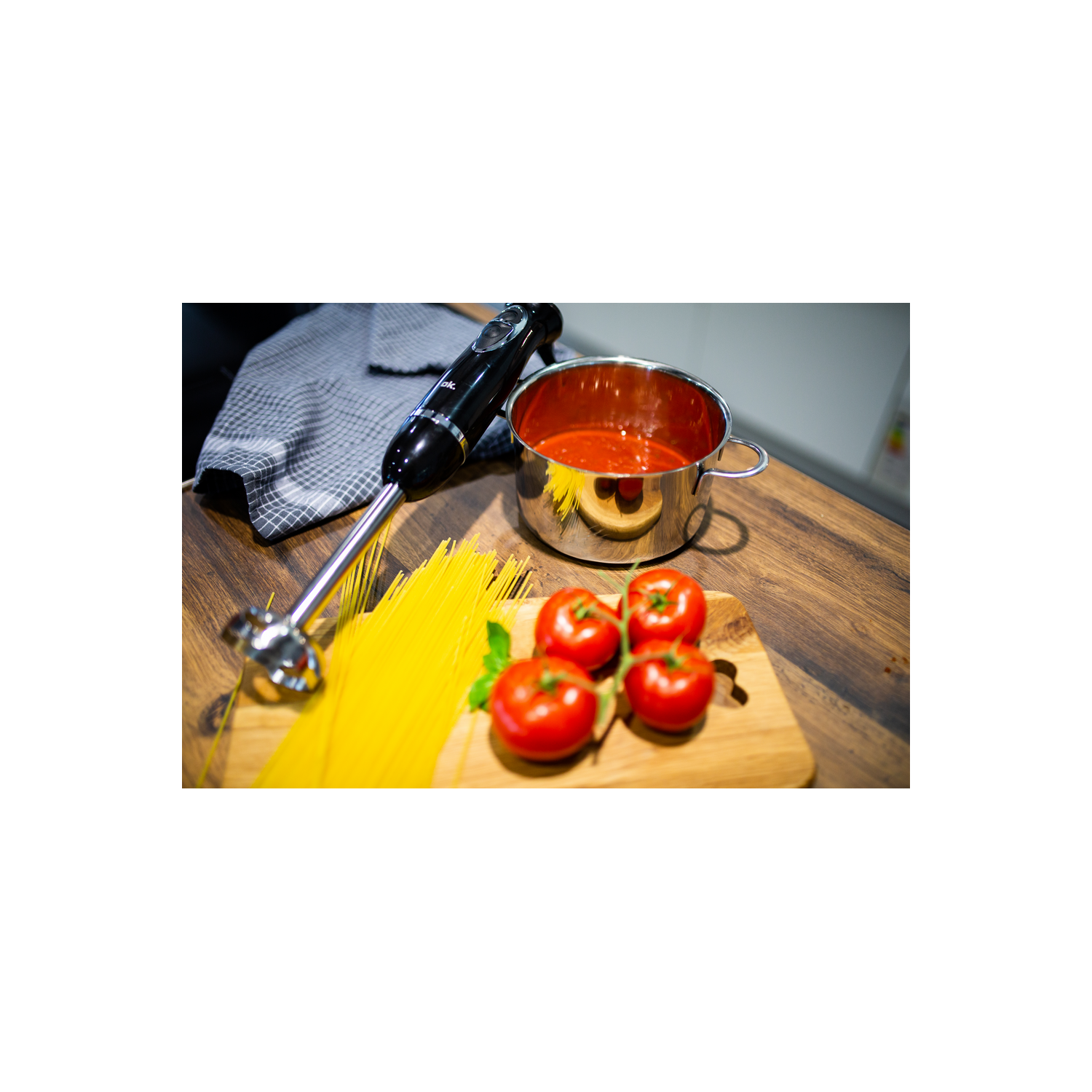 Una batidora de mano negra, una olla de salsa de tomate, espaguetis y tomates en una tabla.
