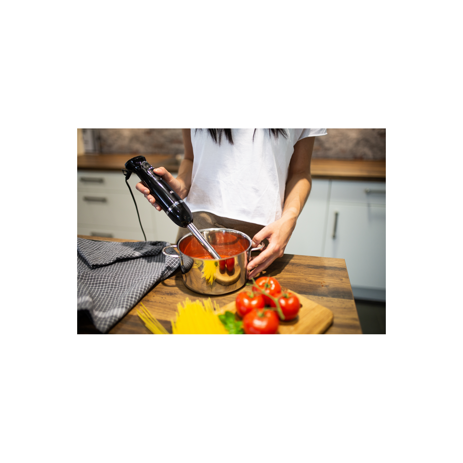 Una persona usando una batidora de inmersión en una olla con salsa de tomate. Tomates y pasta cerca.