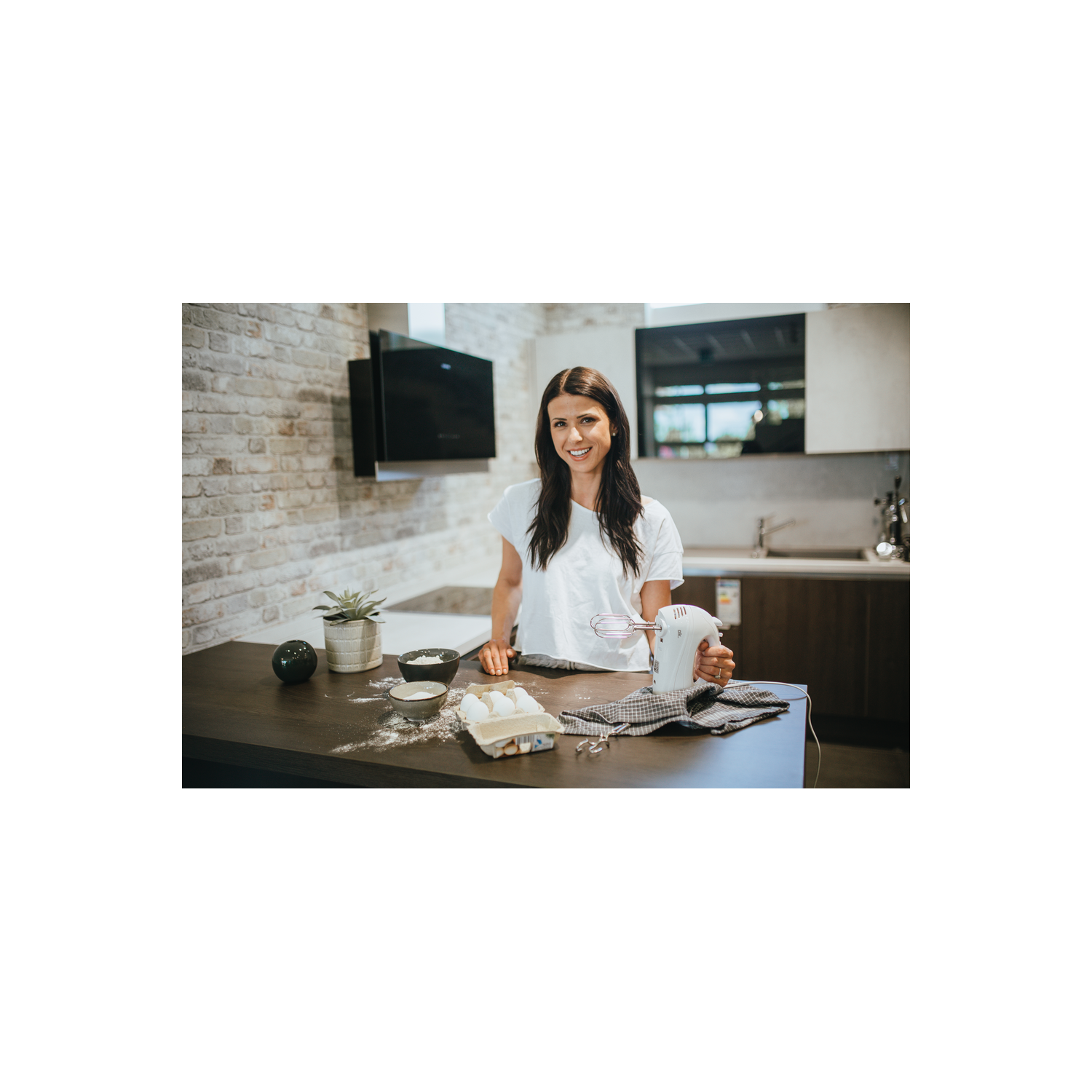 Mujer sonriendo en la cocina, sosteniendo una batidora. Ingredientes en la encimera.