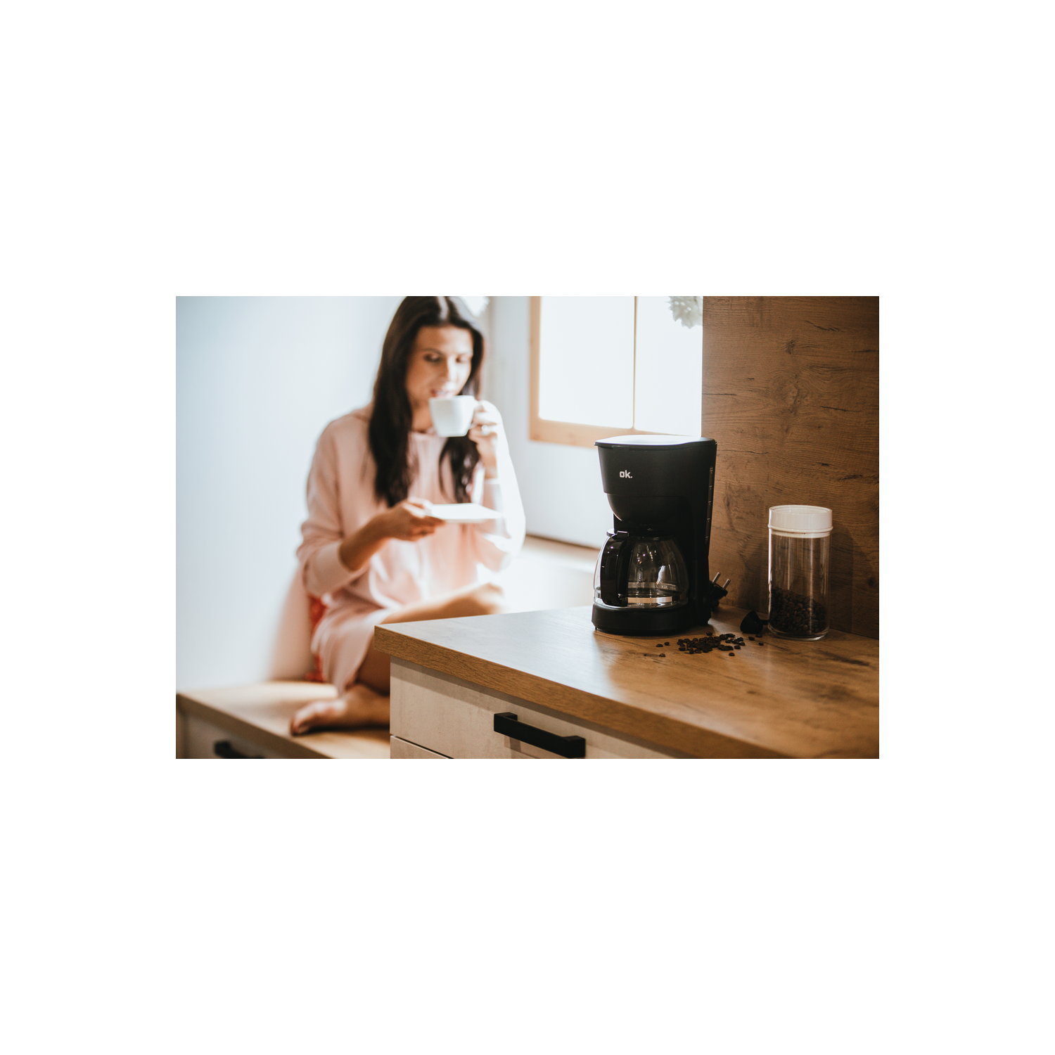 Una cafetera negra sobre una encimera de madera con una mujer sosteniendo una taza.