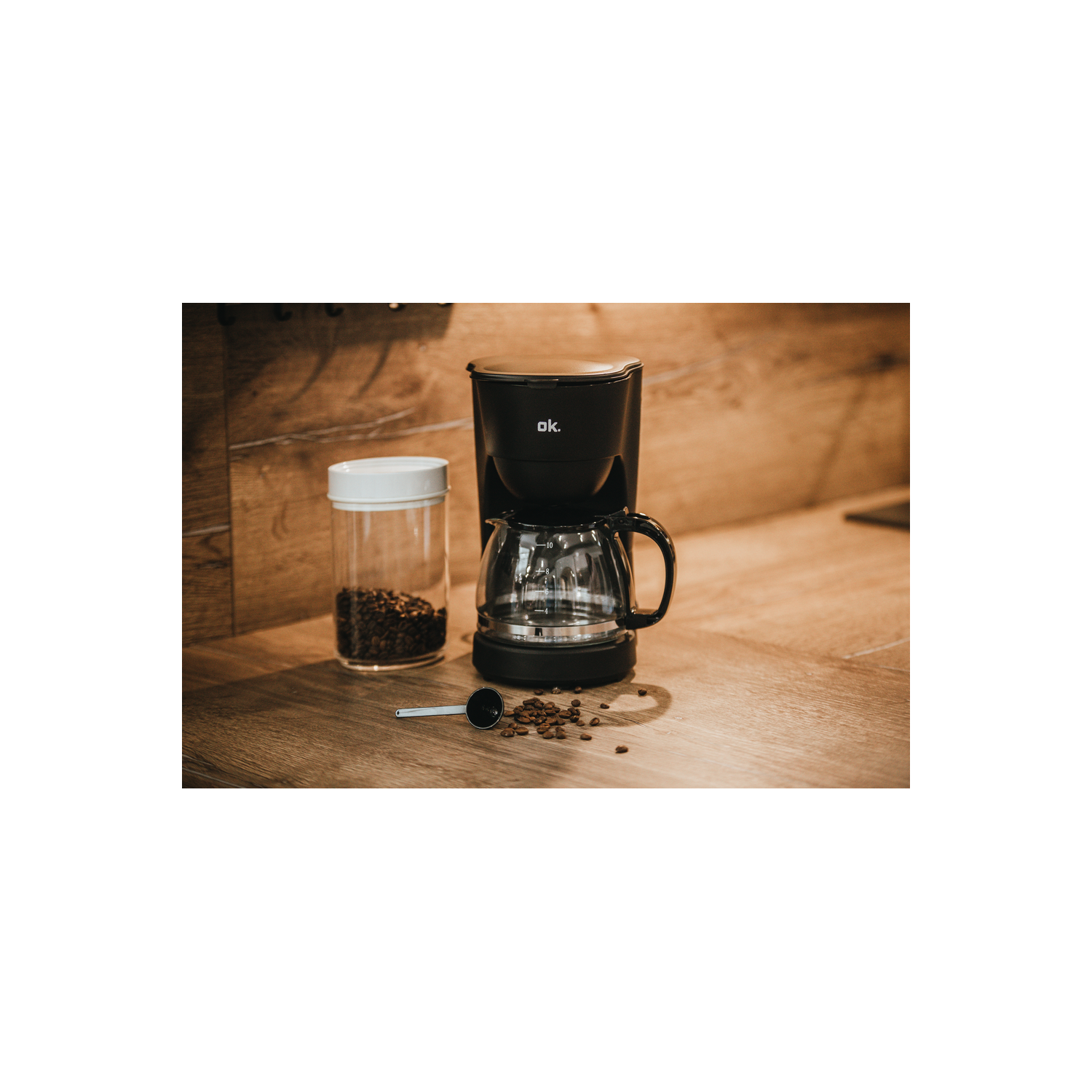 Una cafetera negra sobre una superficie de madera. Un tarro de cristal con granos de café y una cuchara están cerca.