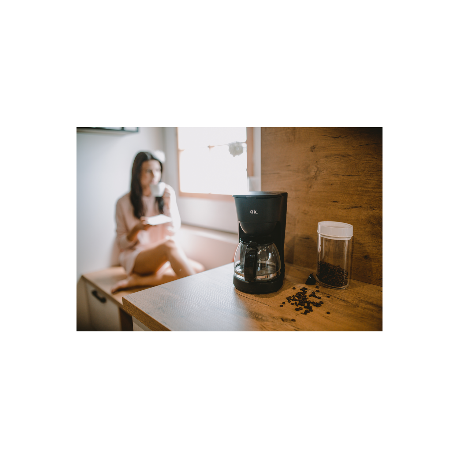 Cafetera en mesa de madera con granos de café y mujer al fondo.
