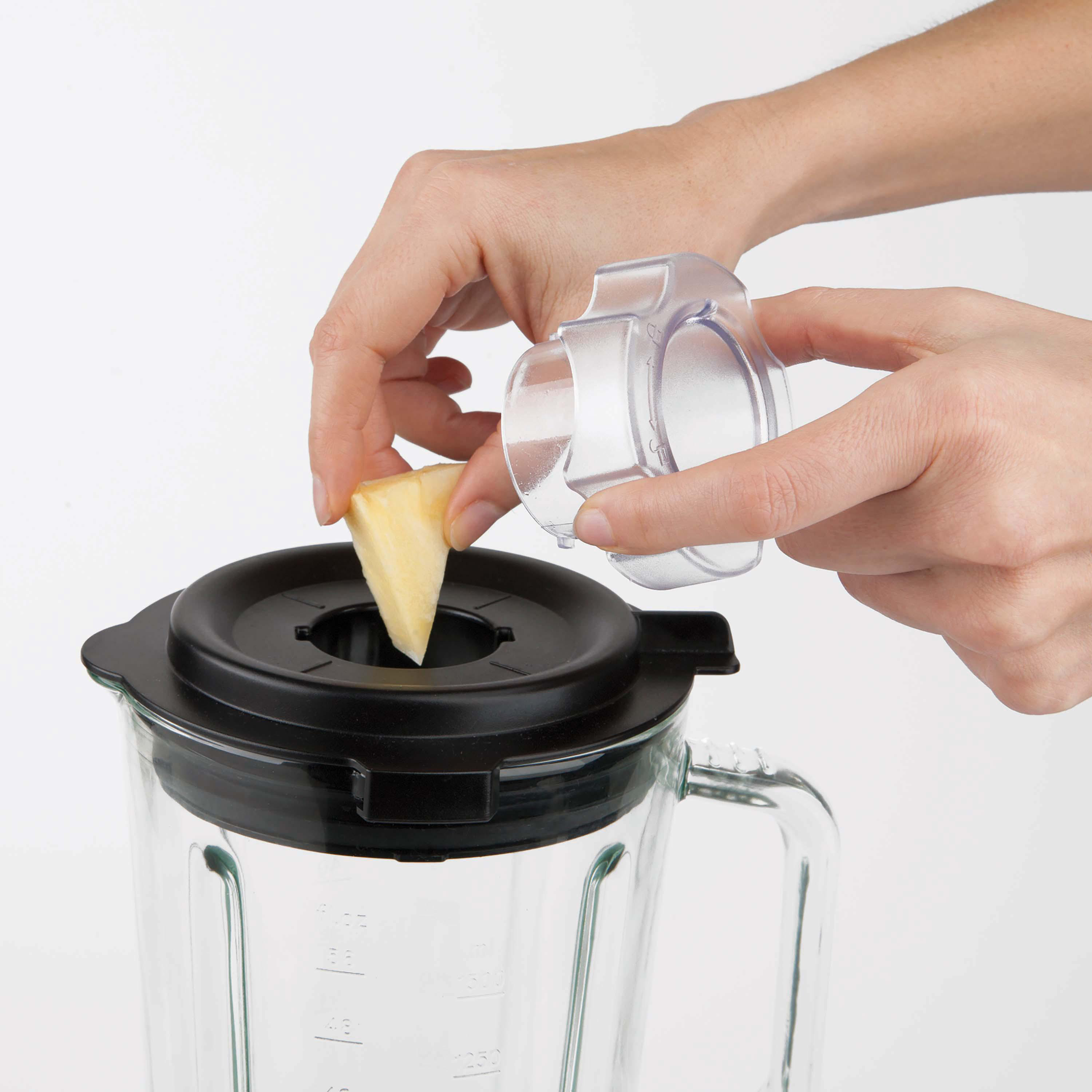 Hände geben Ananasstück in den Mixer. Der Mixer ist aus Glas und hat einen schwarzen Deckel.