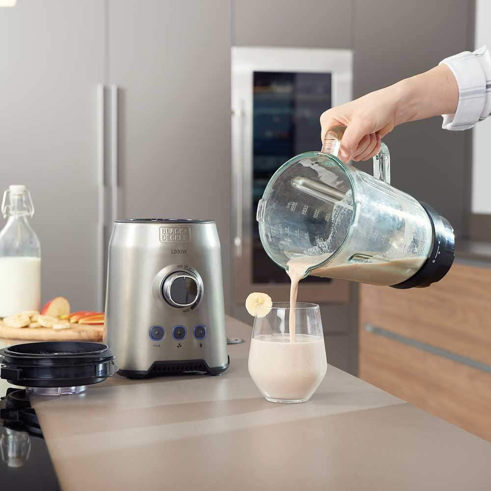 Eine Frau gießt einen Smoothie aus einem Mixer in ein Glas. Aufgeschnittene Banane auf der Theke. Andere Früchte in der Nähe.