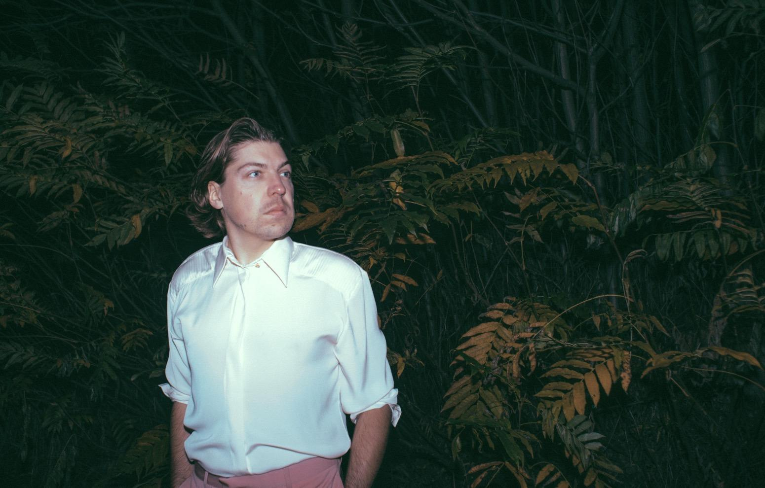 Ein Mann in weißem Hemd und rosa Hose steht inmitten von grünem Laub in einem Wald.