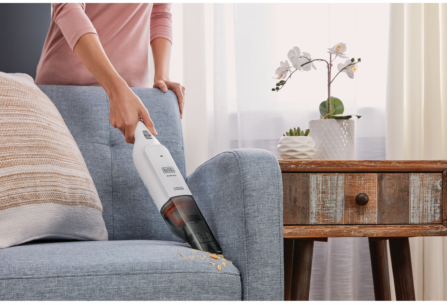 A person using a hand vacuum on a blue sofa, crumbs on the sofa, flowers in the background.